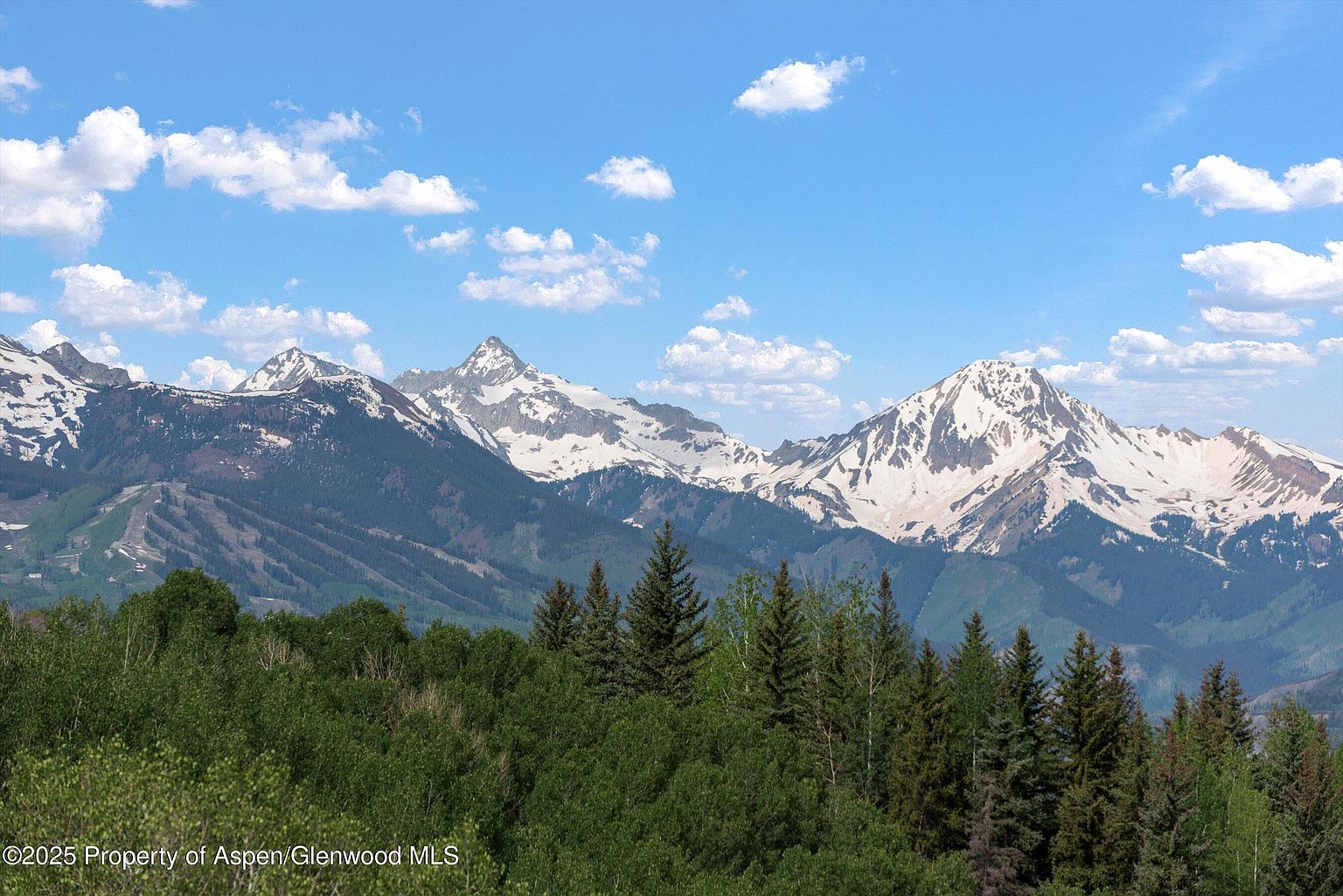 This stunning aerial view showcases a picturesque mountain range with snow-capped peaks under a clear blue sky dotted with fluffy clouds. Lush green trees fill the foreground, creating a beautiful contrast with the rugged mountains. This image would be perfect for highlighting the natural beauty and serene environment of a property.