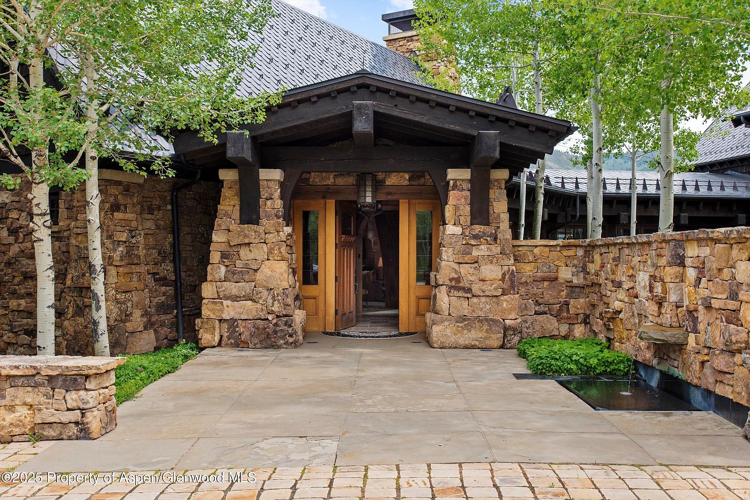 This image showcases the grand entryway of a stone-clad residence. The entrance features a dark wood portico supported by stone pillars, leading to double wooden doors. A small water feature adds a touch of elegance to the paved walkway, complemented by lush greenery. The perspective is a medium shot, emphasizing the architectural details and inviting atmosphere.