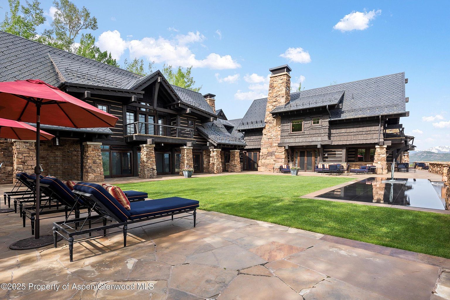This image showcases the rear exterior of a luxurious log home, featuring a sprawling green lawn, a reflecting pool, and a stone patio with lounge chairs. The architectural style emphasizes natural materials like wood and stone, creating a rustic yet elegant aesthetic. The view suggests a private and serene outdoor living space, perfect for relaxation and entertainment.