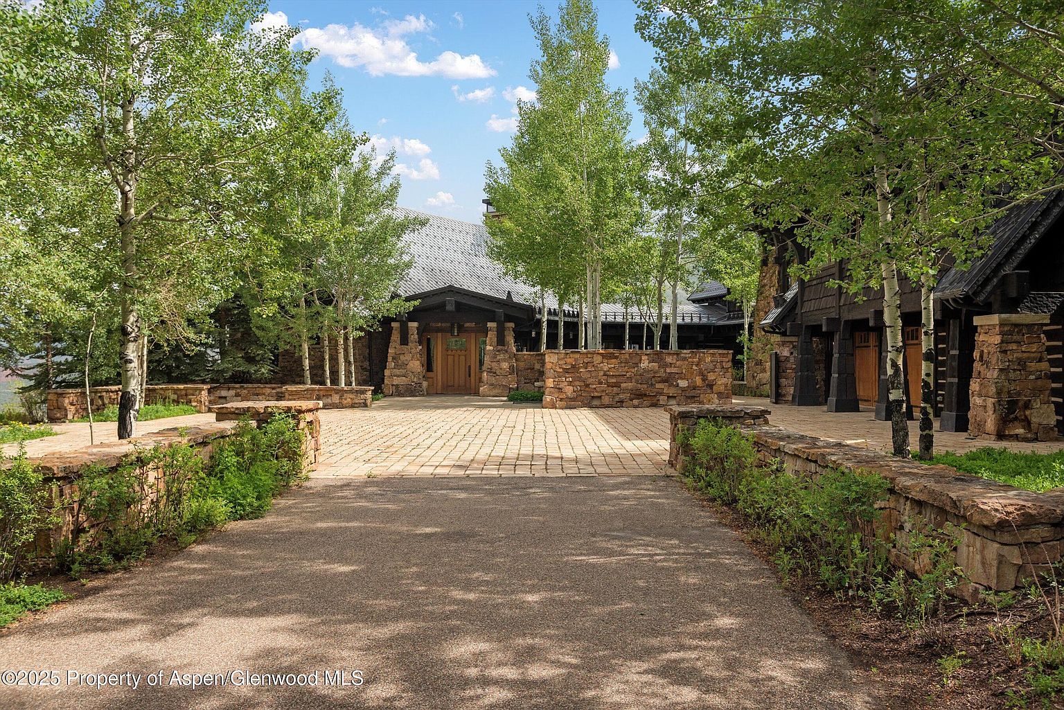 This image showcases the grand entryway of a luxurious home, featuring a stone-paved driveway leading to a stone facade with a wooden door. Mature trees frame the entrance, adding to the property's curb appeal and sense of privacy. The architecture blends natural materials with a sophisticated design, creating an inviting and impressive arrival experience.