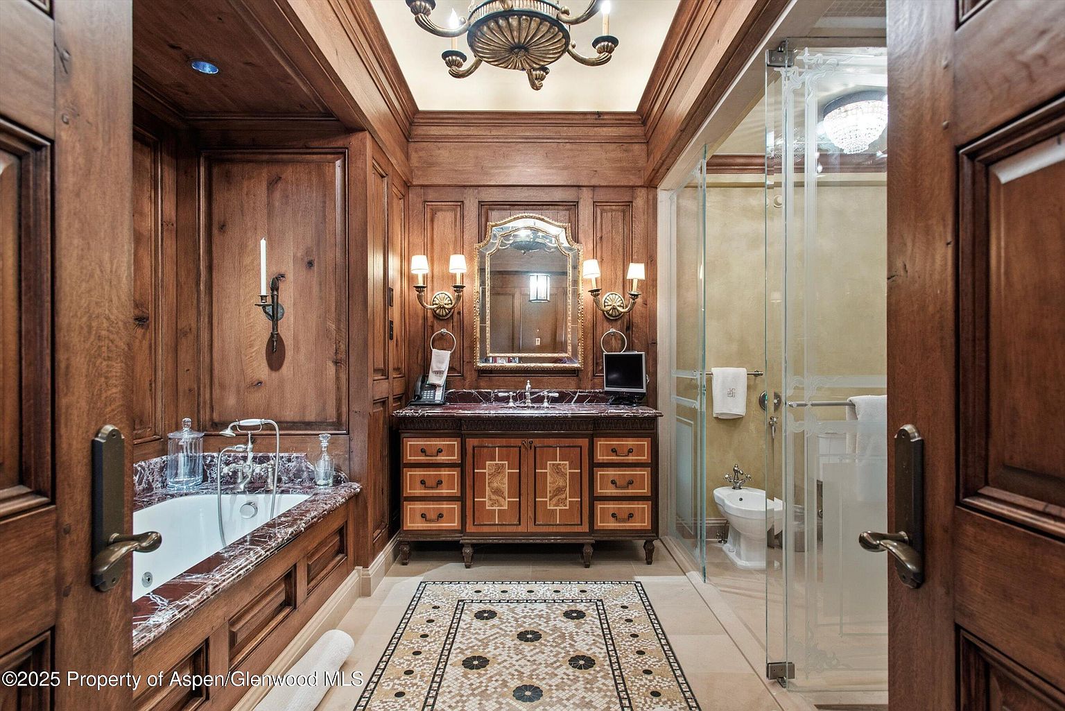 This is a luxurious primary bathroom featuring rich wood paneling, a marble-topped vanity with a decorative mirror and sconces, and a separate shower enclosure. The floor is adorned with an intricate mosaic tile pattern, adding to the room's opulent feel. A soaking tub is visible to the left, suggesting a relaxing and spa-like atmosphere.