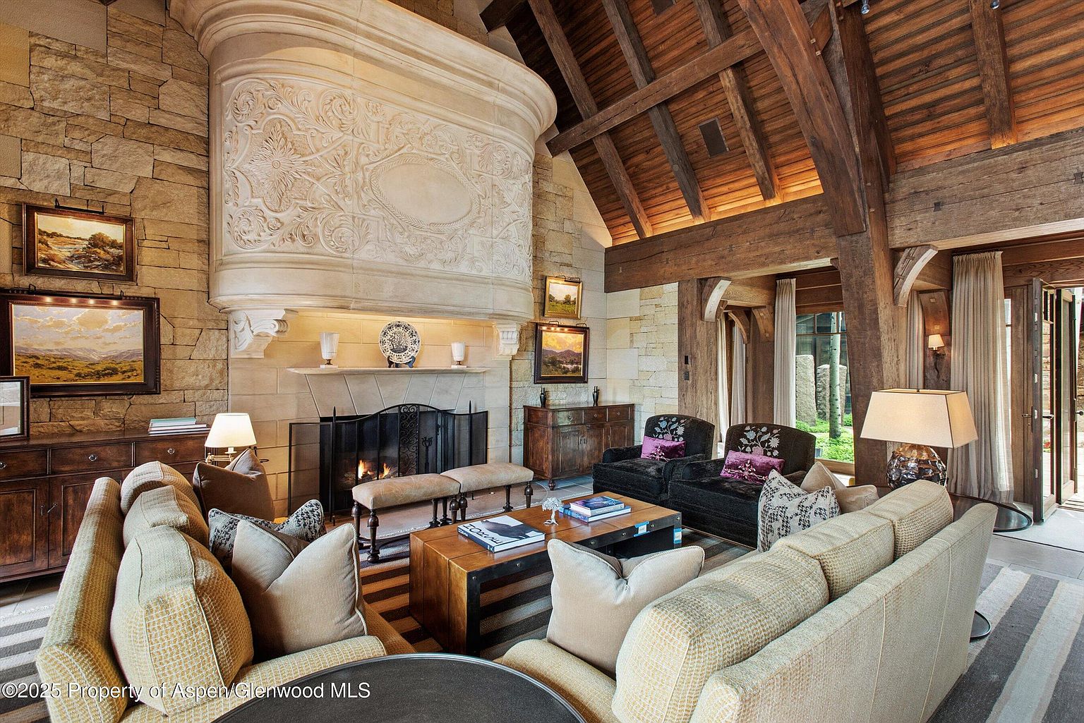 This is a luxurious living room featuring a grand stone fireplace with intricate detailing, complemented by exposed wooden beams on the ceiling. The room is furnished with comfortable sofas and armchairs, arranged around a wooden coffee table on a striped rug. The space exudes warmth and sophistication, perfect for relaxation and entertaining.