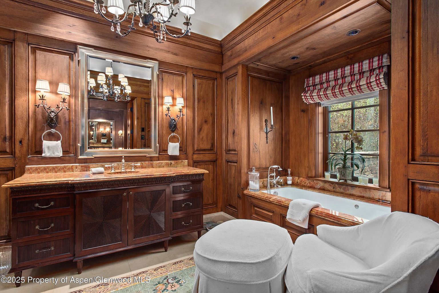 This is a luxurious primary bathroom featuring rich wood paneling on the walls and ceiling, creating a warm and sophisticated ambiance. A marble-topped vanity with a large mirror and sconces is visible, along with a soaking tub positioned by a window with a red and white striped shade. The room is furnished with a white chair and ottoman, adding to the spa-like feel.