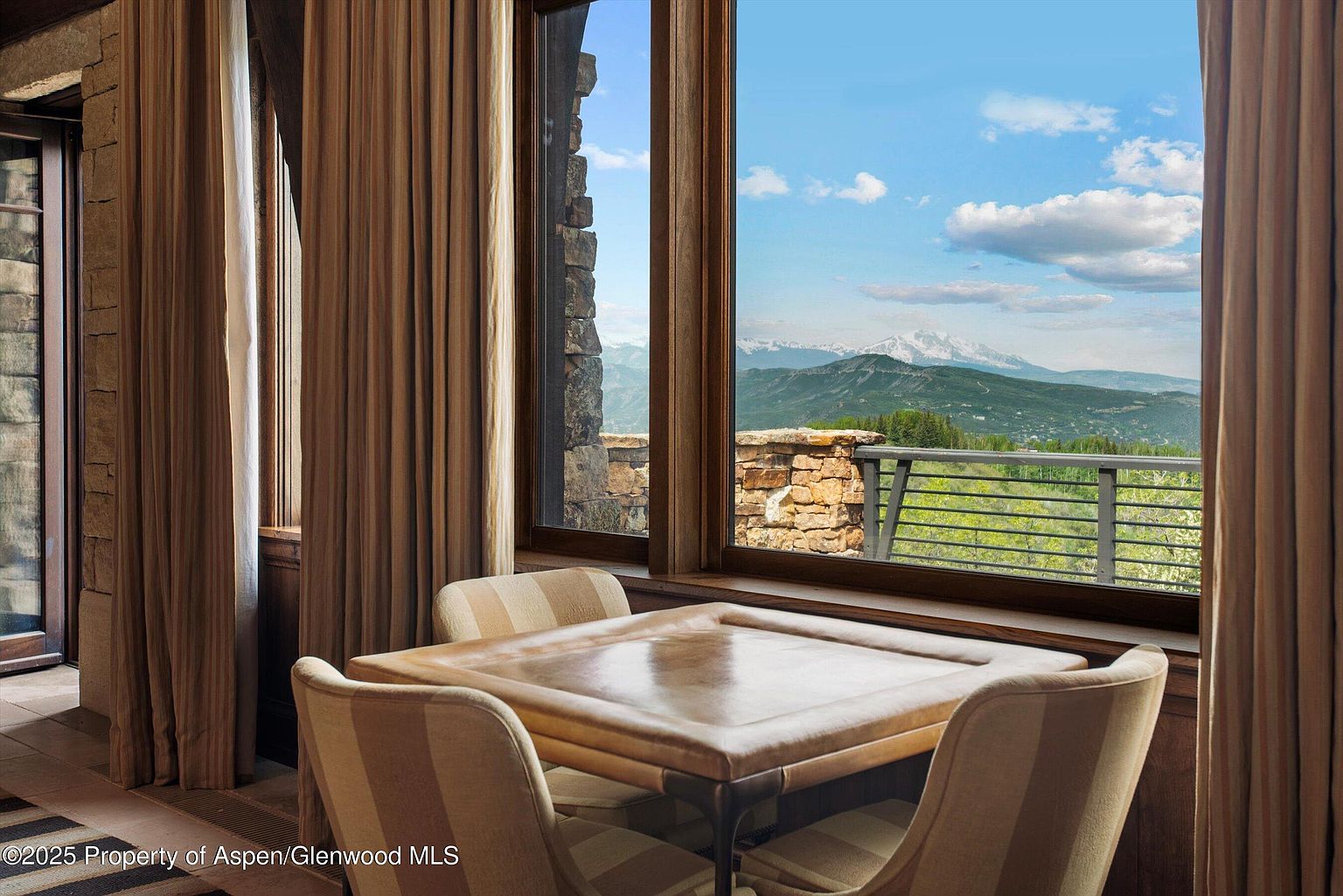 This interior shot showcases a dining area with a square table and four chairs, positioned in front of a large window offering a scenic mountain view. The room is decorated with neutral-toned curtains and stone accents, creating a warm and inviting atmosphere. The space appears well-lit and designed to maximize the natural light and picturesque landscape.