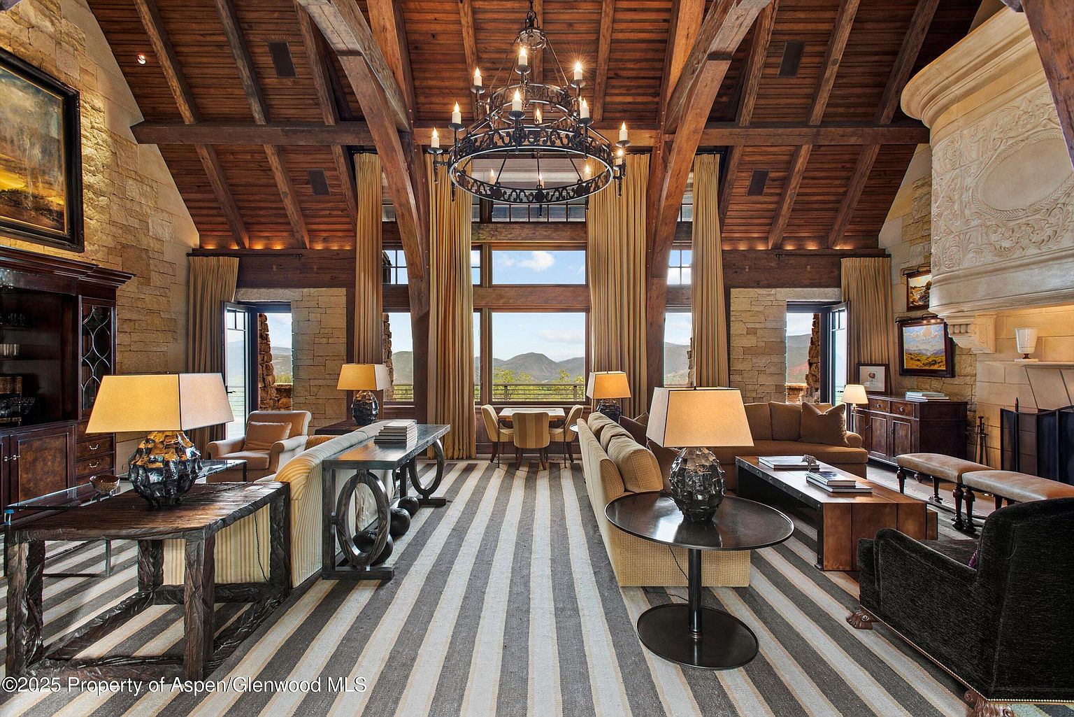 This grand living room features a high, beamed ceiling and a striking chandelier, creating a luxurious and spacious atmosphere. Large windows offer scenic mountain views, complemented by elegant drapery. The room is furnished with comfortable seating arrangements and stylish tables, all set upon a striped rug that adds a touch of modern sophistication.