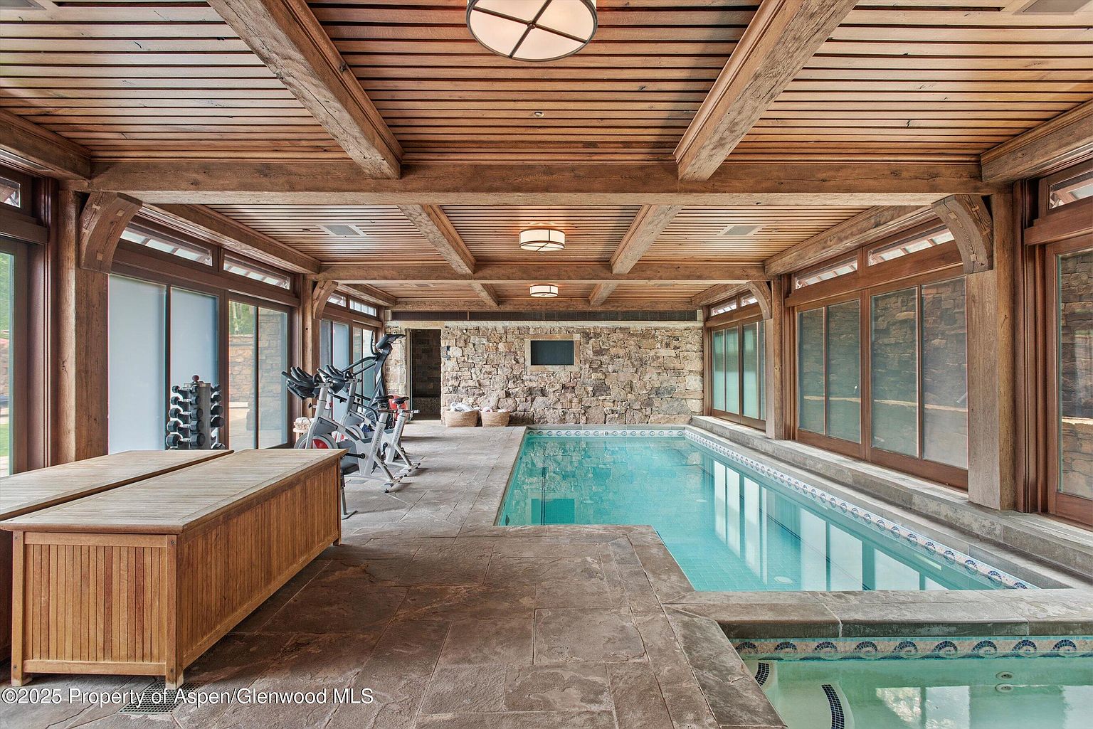 This interior shot showcases a luxurious home gym featuring an indoor pool and workout area. The space is characterized by a wooden beamed ceiling, stone accent wall, and large windows providing ample natural light. Exercise equipment is visible alongside the pool, creating a combined fitness and relaxation environment.