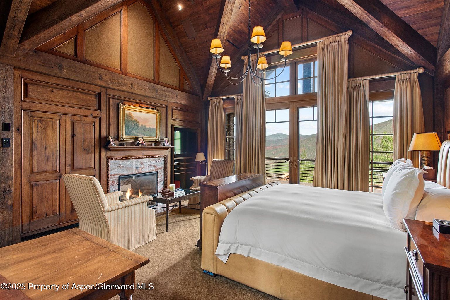 This is a luxurious primary bedroom featuring a grand fireplace, rich wood paneling, and a vaulted ceiling with exposed beams. Large windows offer stunning mountain views, complemented by elegant drapery and a sophisticated chandelier. The room exudes warmth and comfort, making it a perfect retreat.