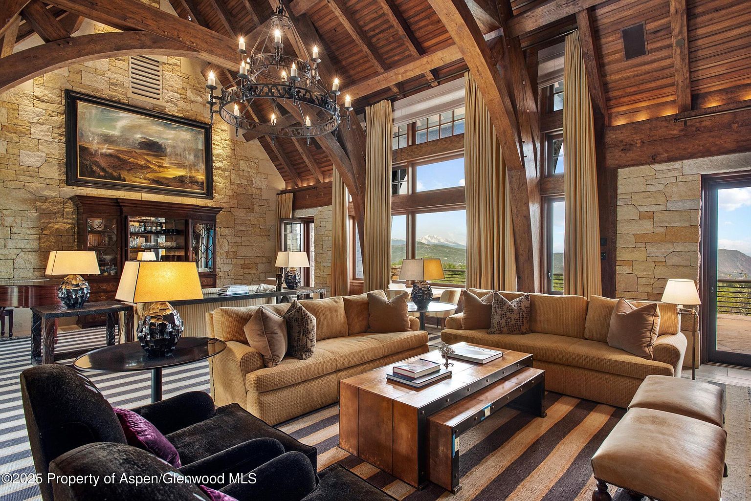 This is a luxurious living room featuring a high vaulted ceiling with exposed wooden beams and a stone accent wall. The room is furnished with comfortable sofas, a wooden coffee table, and a striped rug. Large windows offer a scenic view of the mountains, and a grand chandelier hangs from the ceiling, adding to the room's elegant ambiance.