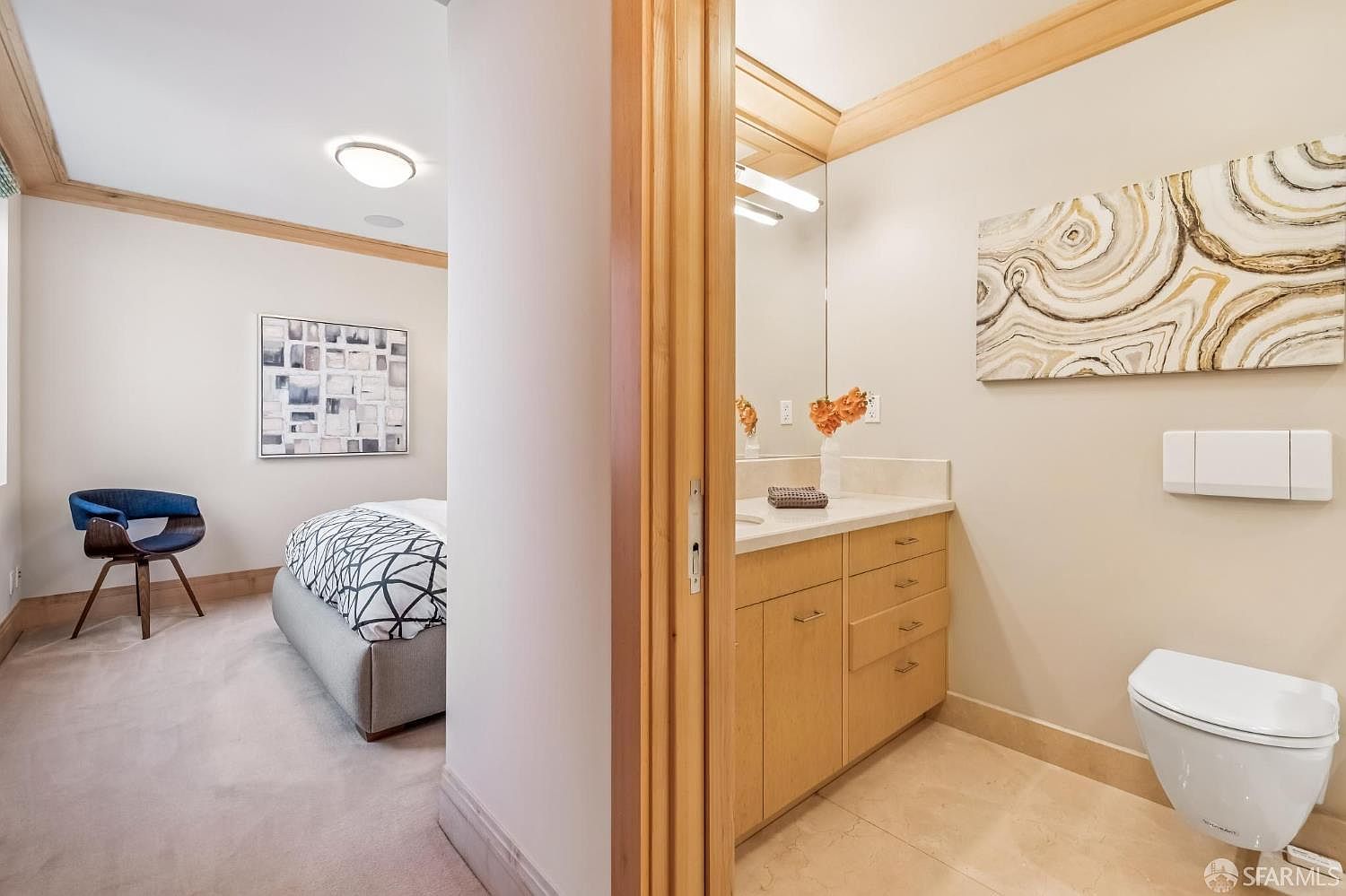 This image captures a view from a bedroom into an adjacent bathroom, showcasing a seamless transition between living and utility spaces. The bathroom features light wood cabinetry, a neutral stone countertop, and a wall-mounted toilet, while the bedroom is furnished with a modern bed and a blue accent chair. The neutral color palette and clean lines create a cohesive and contemporary aesthetic throughout both rooms.