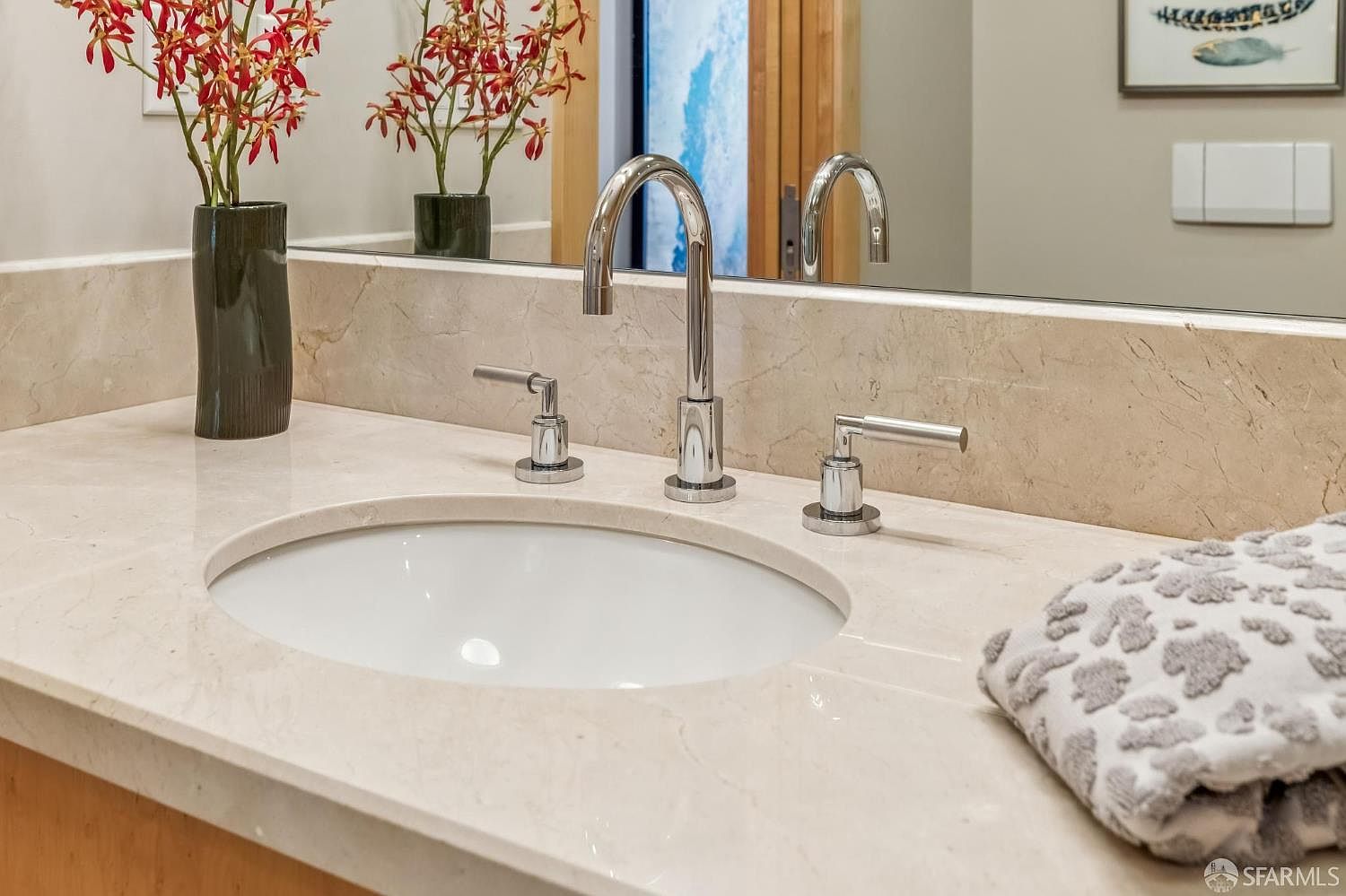 This close-up shot captures a clean, modern bathroom vanity featuring a light-toned stone countertop and an undermount sink. A sleek, polished chrome faucet is centered between two matching handles, complemented by a decorative vase of red flowers and a soft, patterned hand towel resting on the side. The scene is reflected in a large mirror, creating a bright and polished aesthetic suitable for a contemporary home.
