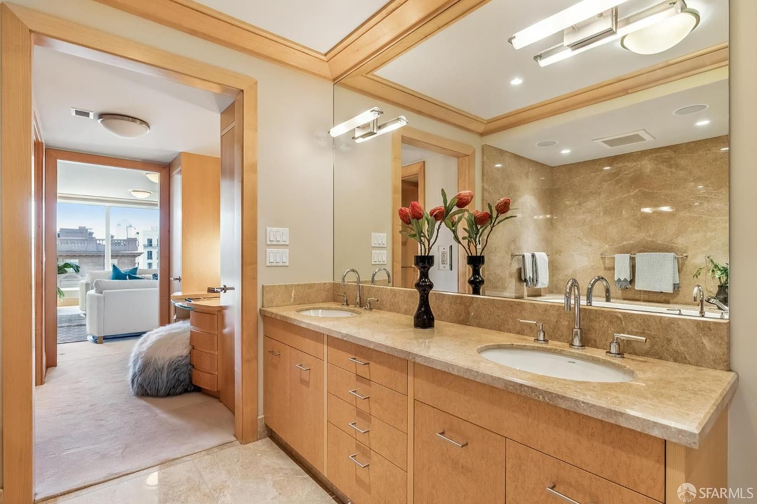 This primary bathroom features a dual-sink vanity with light wood cabinetry and a neutral stone countertop. A large, wall-to-wall mirror reflects the space, including a glimpse into an adjacent room with a view of the outdoors. The room is accented by decorative floral arrangements and modern lighting fixtures, creating a bright and polished atmosphere.