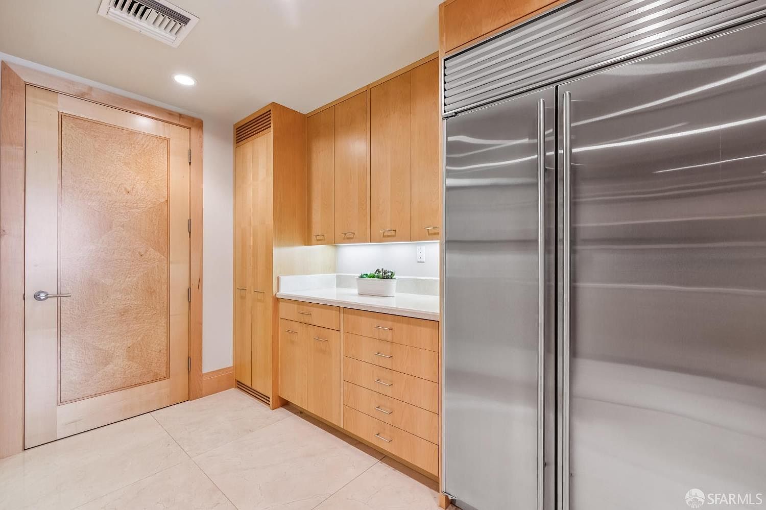 This kitchen area features light wood cabinetry with a clean, minimalist aesthetic and a large stainless steel refrigerator. A white countertop provides a workspace with under-cabinet lighting, while a tall, matching wood-paneled door stands to the left. The space is finished with light-colored tile flooring, creating a bright and organized atmosphere.
