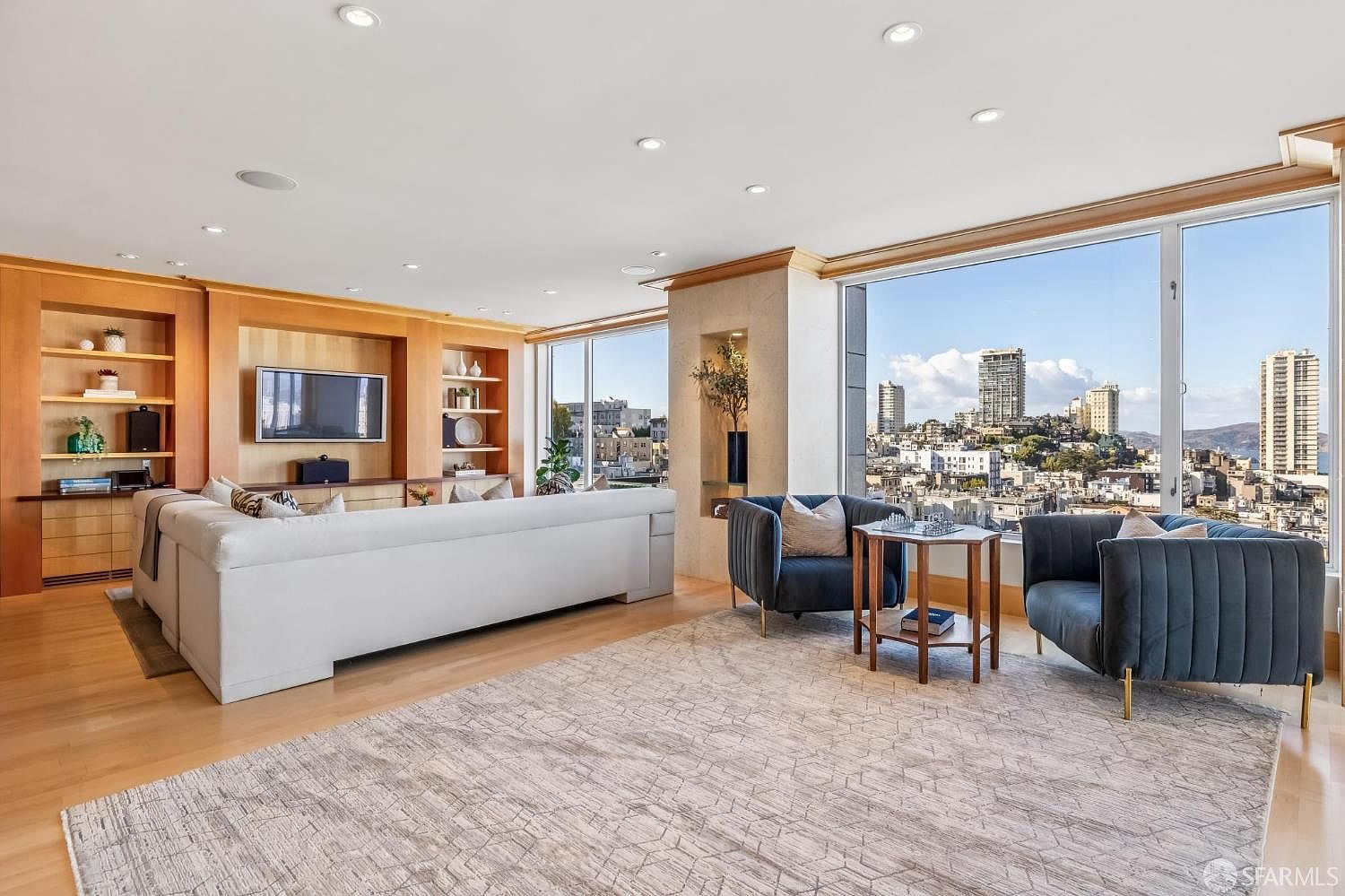 This spacious, light-filled living room features a large, neutral-toned sectional sofa and two dark blue velvet armchairs arranged around a central wooden side table. The room is anchored by a large, textured area rug over light wood flooring and includes custom built-in shelving with integrated lighting. Expansive windows provide a panoramic view of the city skyline, creating a bright and sophisticated atmosphere.