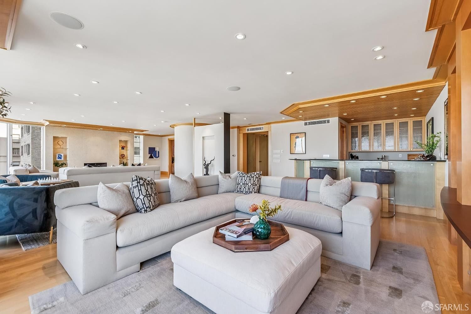 This spacious, open-concept living area features a large, light-toned sectional sofa arranged around a central ottoman with a wooden tray. The room is illuminated by numerous recessed ceiling lights and flows seamlessly into a bar area with glass-front cabinetry and bar seating. The neutral color palette and light wood flooring create a bright, airy, and sophisticated atmosphere suitable for entertaining.
