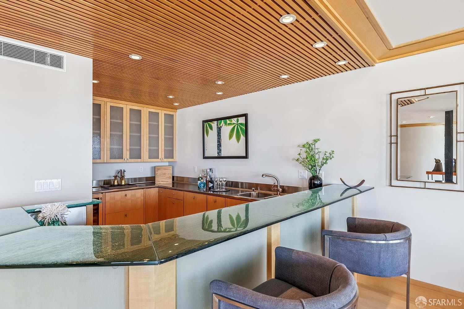 This sophisticated wet bar area features a sleek, glass-topped counter with two modern, upholstered armchairs, perfect for entertaining. The space is defined by warm wood cabinetry, a dark stone countertop, and a distinctive slatted wood ceiling with recessed lighting. A framed botanical print and a decorative mirror add elegant touches to the clean, neutral walls, creating a refined and inviting atmosphere.