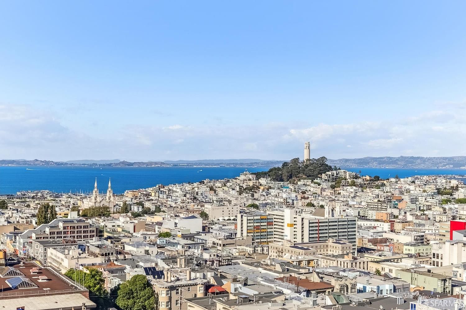 This high-angle aerial view captures a sprawling cityscape of San Francisco, featuring dense urban architecture and iconic landmarks like Coit Tower in the distance. The scene showcases a mix of residential and commercial buildings under a clear blue sky, with the sparkling waters of the bay visible in the background. The perspective provides a comprehensive look at the neighborhood's density and its proximity to the waterfront.