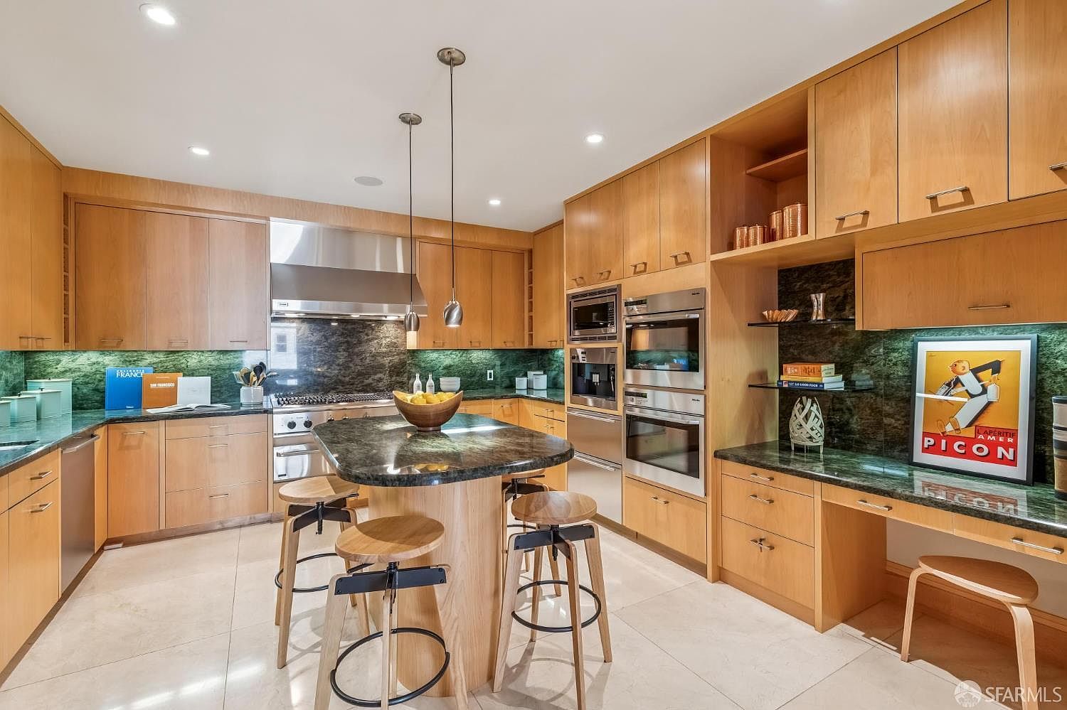 This modern kitchen features light wood cabinetry, a central island with a stone countertop, and high-end stainless steel appliances including a built-in coffee system and double ovens. A green stone backsplash adds a vibrant pop of color, complemented by a dedicated desk area and a framed vintage poster on the wall. The space is illuminated by recessed lighting and two sleek pendant lights hanging over the island, creating a warm and functional culinary environment.