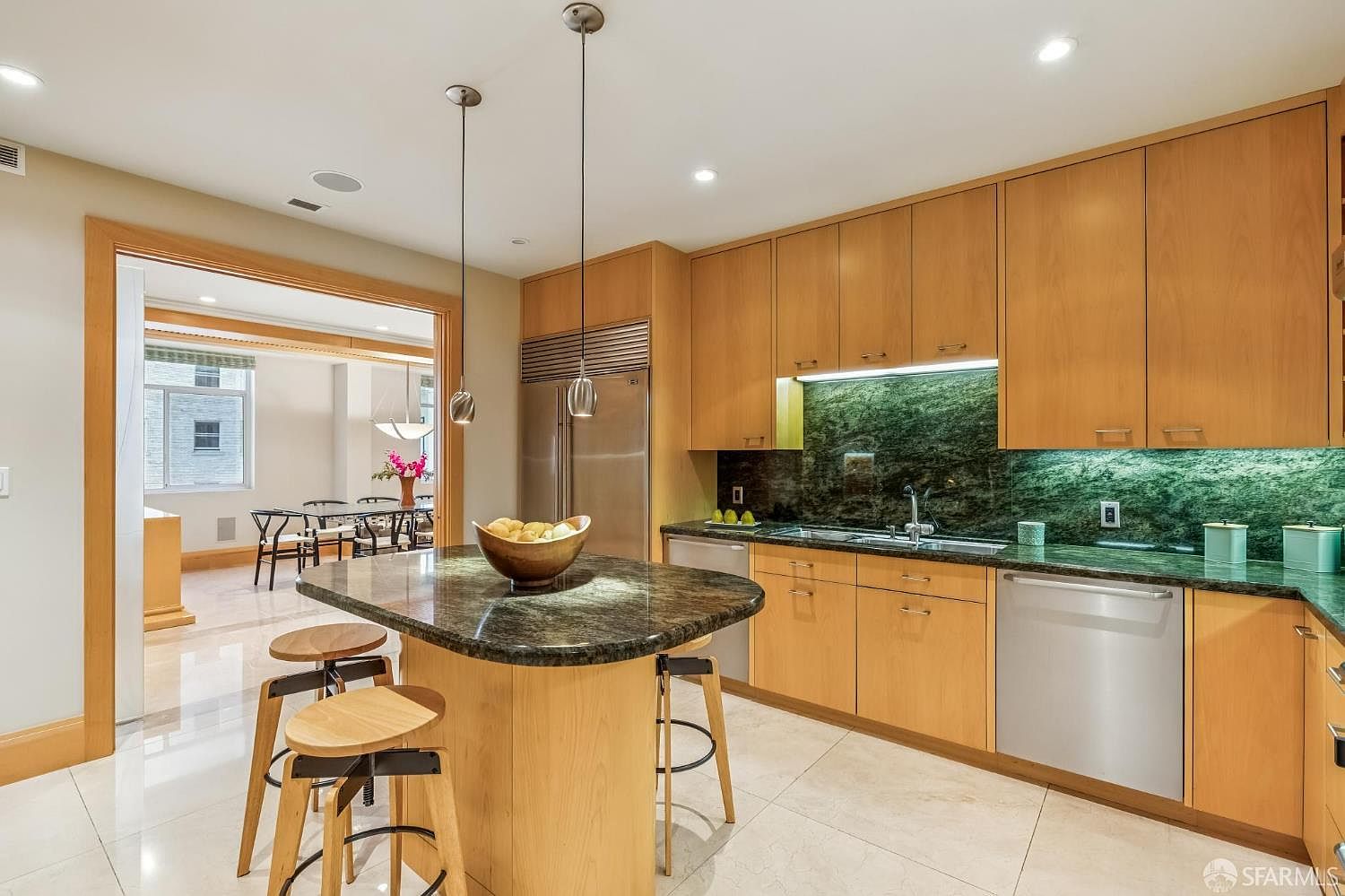 This modern kitchen features light wood cabinetry paired with dark, patterned stone countertops and a matching backsplash. A central island provides additional workspace and seating with two stools, illuminated by two sleek pendant lights. The space is bright and open, with a clear view through a wide doorway into an adjacent dining area, creating a cohesive and airy floor plan.