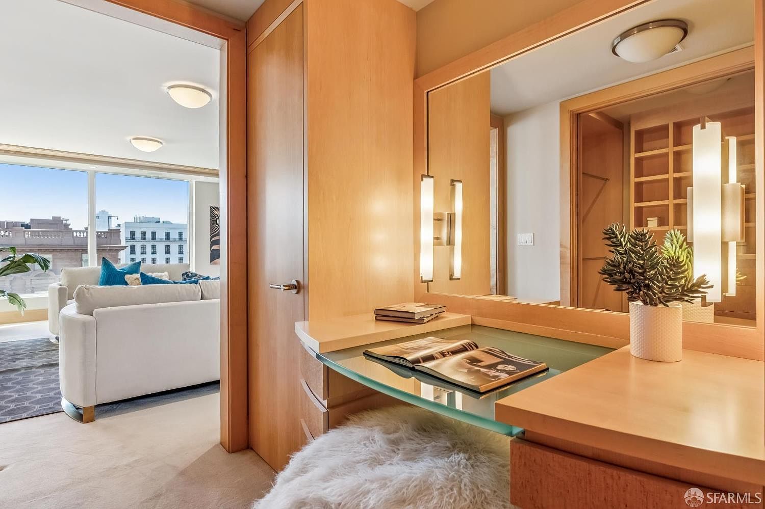 This image captures a sophisticated dressing area featuring a built-in vanity with a glass top and light wood cabinetry. A large mirror reflects the adjacent walk-in closet space, while a doorway provides a clear view into a bright, modern living room with large windows overlooking the city. The combination of warm wood tones, soft textures like the faux-fur stool, and the seamless transition between rooms creates an elegant and functional atmosphere.