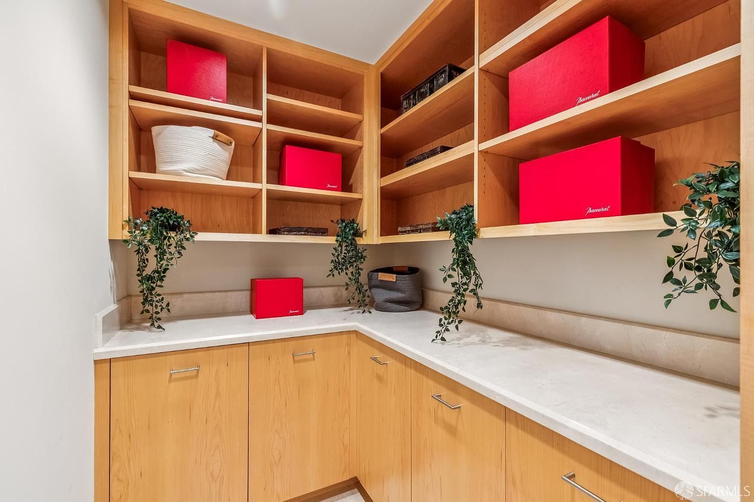 This organized walk-in closet features light wood cabinetry with ample open shelving and lower storage cabinets topped with a light-colored stone counter. Several bright red Baccarat boxes are displayed on the shelves, adding a pop of color to the neutral space. Decorative hanging plants are placed throughout, providing a touch of greenery to the clean, functional storage area.