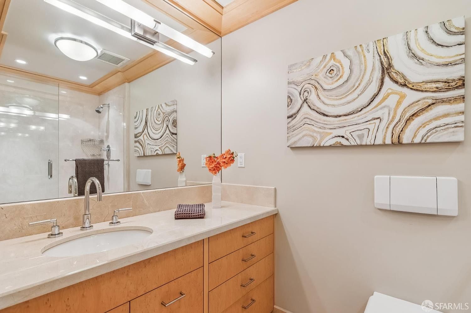 This bright and well-maintained bathroom features a light wood vanity with a stone countertop and an integrated sink. A large mirror spans the wall above the vanity, reflecting a glass-enclosed shower and a piece of abstract wall art. The space is accented by warm lighting and a clean, neutral color palette, creating a polished and functional atmosphere.