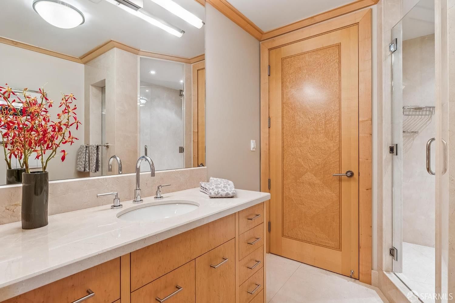 This primary bathroom features a light wood vanity with a stone countertop and a single undermount sink. A large mirror spans the wall above the vanity, reflecting the bright lighting and a glass-enclosed shower stall to the right. The space is finished with neutral-toned wall tiles and a light wood door, creating a clean and cohesive aesthetic.