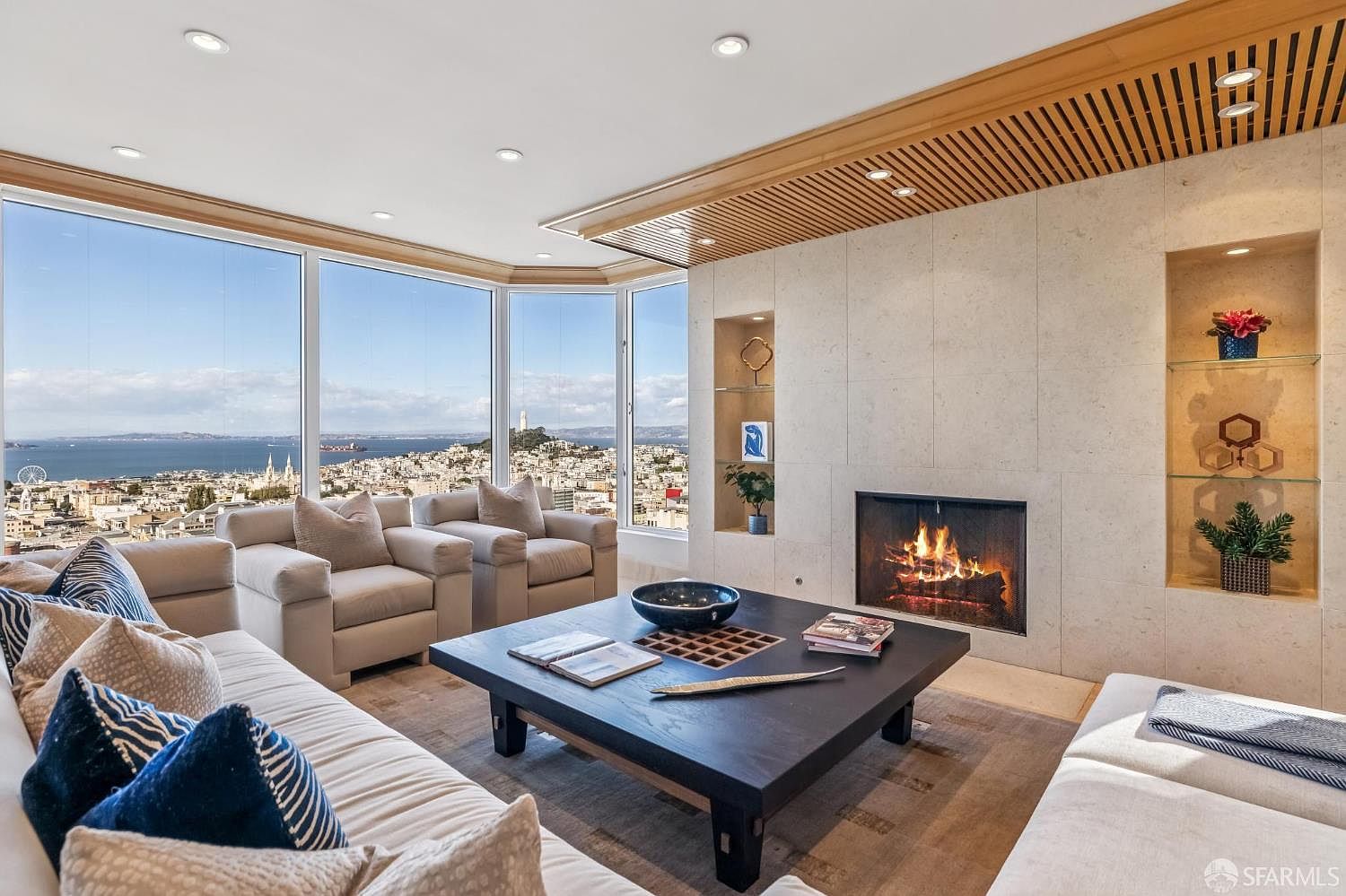 This sophisticated living room features a large, dark wood coffee table as a central focal point, surrounded by plush, light-toned seating. A prominent stone-clad chimney breast houses a warm, glowing fireplace, flanked by recessed shelving units displaying decorative items. Expansive windows provide a panoramic view of the city skyline and bay, creating a bright and airy atmosphere enhanced by a slatted wood ceiling detail.