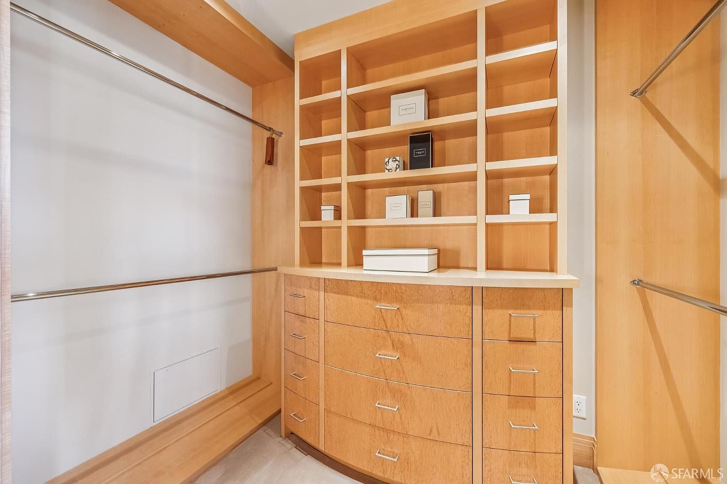 This organized walk-in closet features custom light wood cabinetry with a combination of open shelving and multiple drawers. The space includes dual-level hanging rods on either side, providing ample storage for clothing and accessories. The neutral, clean aesthetic creates a functional and well-lit storage area.