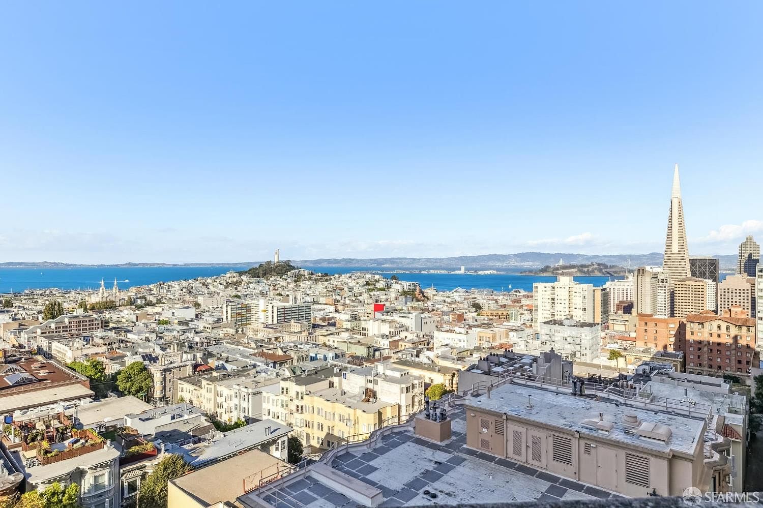This high-angle aerial view captures a sweeping cityscape of San Francisco, showcasing the iconic Transamerica Pyramid and the surrounding urban density. The perspective looks out over a mix of residential rooftops and commercial buildings toward the sparkling blue waters of the bay and distant hills. The clear, bright sky emphasizes the expansive nature of the view, highlighting the property's prime location within the city.