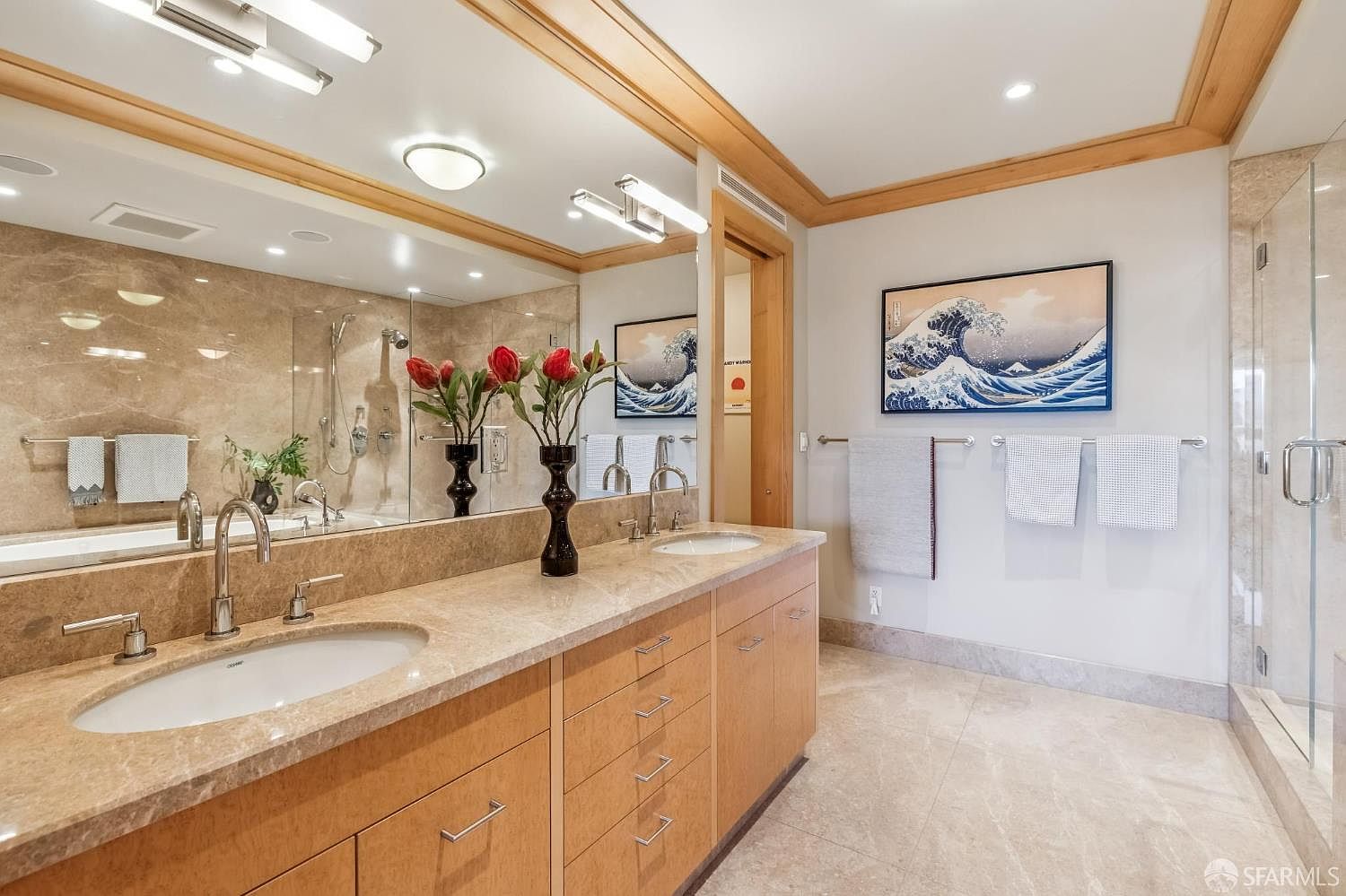 This spacious primary bathroom features a double vanity with light wood cabinetry and a neutral stone countertop. A large, wall-spanning mirror reflects the room, including a glass-enclosed shower and a framed print of 'The Great Wave off Kanagawa' hanging on the wall. The space is finished with light-toned stone flooring and walls, creating a bright and cohesive aesthetic.