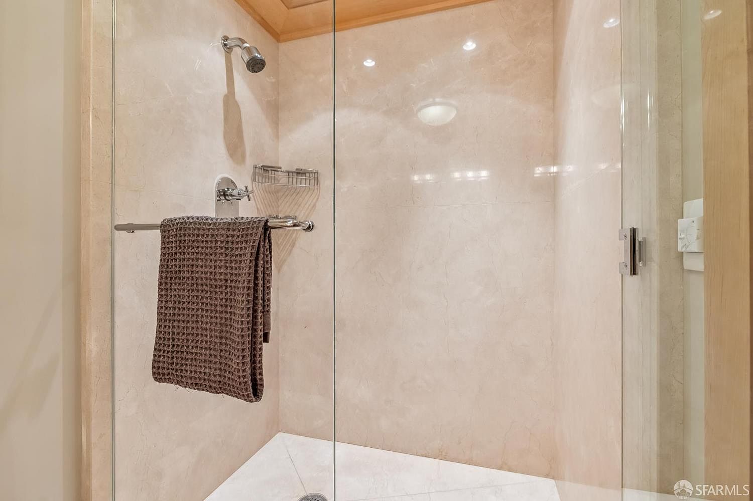 This image captures a walk-in shower enclosure featuring light-toned stone wall tiles and a clean white floor. A dark, textured towel hangs over the chrome handle of the glass door, while a metal caddy is mounted on the wall for storage. The perspective is a direct, eye-level view looking into the shower stall, highlighting the minimalist and functional design of the space.