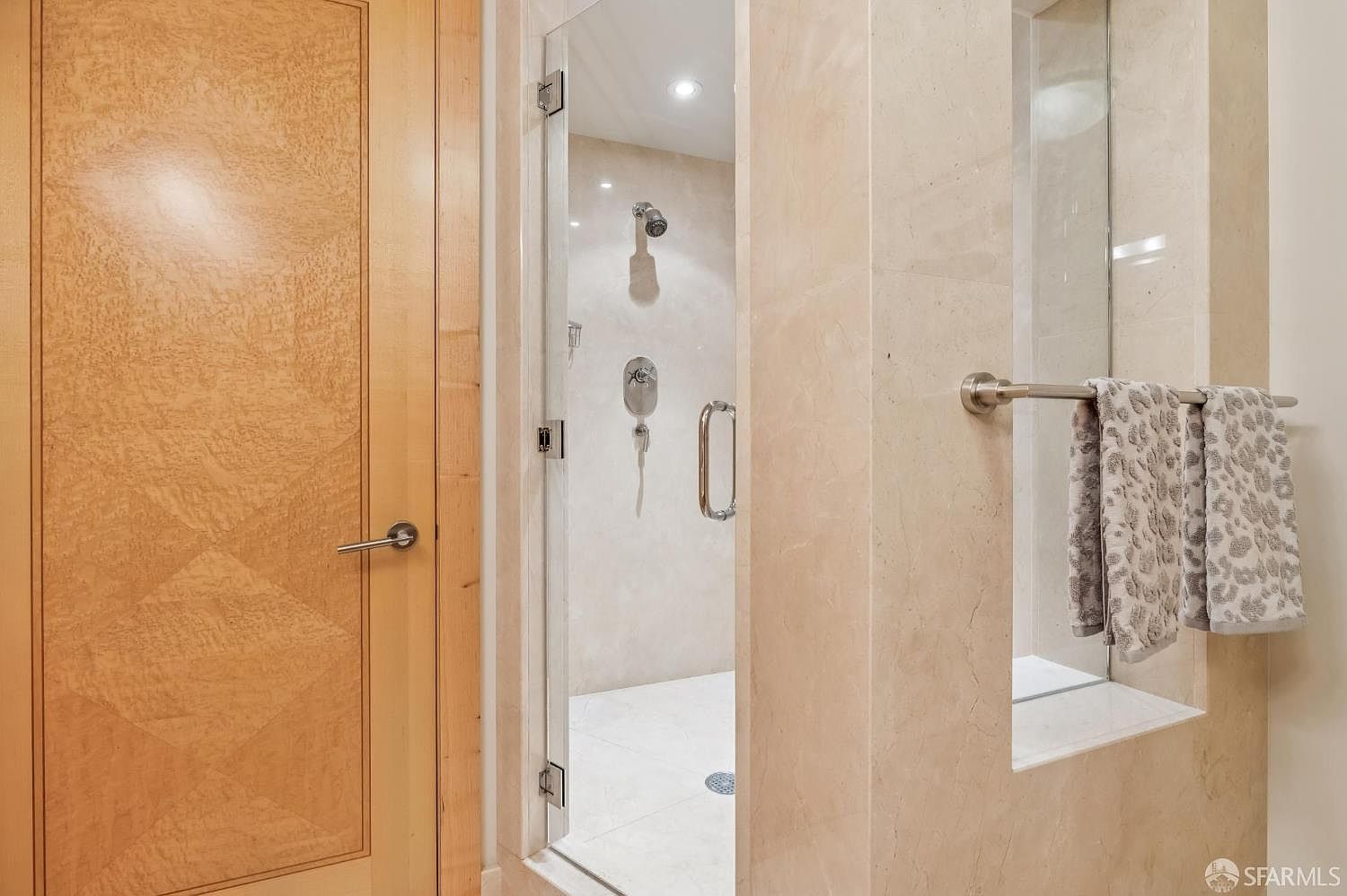 This image captures a modern bathroom space featuring a glass-enclosed walk-in shower with light-toned stone wall cladding. To the left, a light wood door with a geometric panel design adds warmth, while a recessed wall niche on the right provides convenient storage for towels on a metal rack. The perspective is centered on the shower entrance, highlighting the clean lines and neutral color palette of the contemporary design.