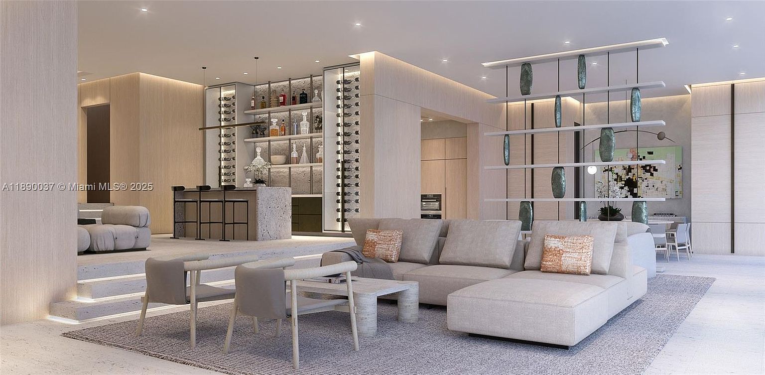 This is a spacious and modern living room featuring a large sectional sofa with decorative pillows, a unique coffee table, and two chairs. The room is decorated in neutral tones with a contemporary art installation and a bar area in the background. The perspective is wide, showcasing the open floor plan and luxurious feel of the space.