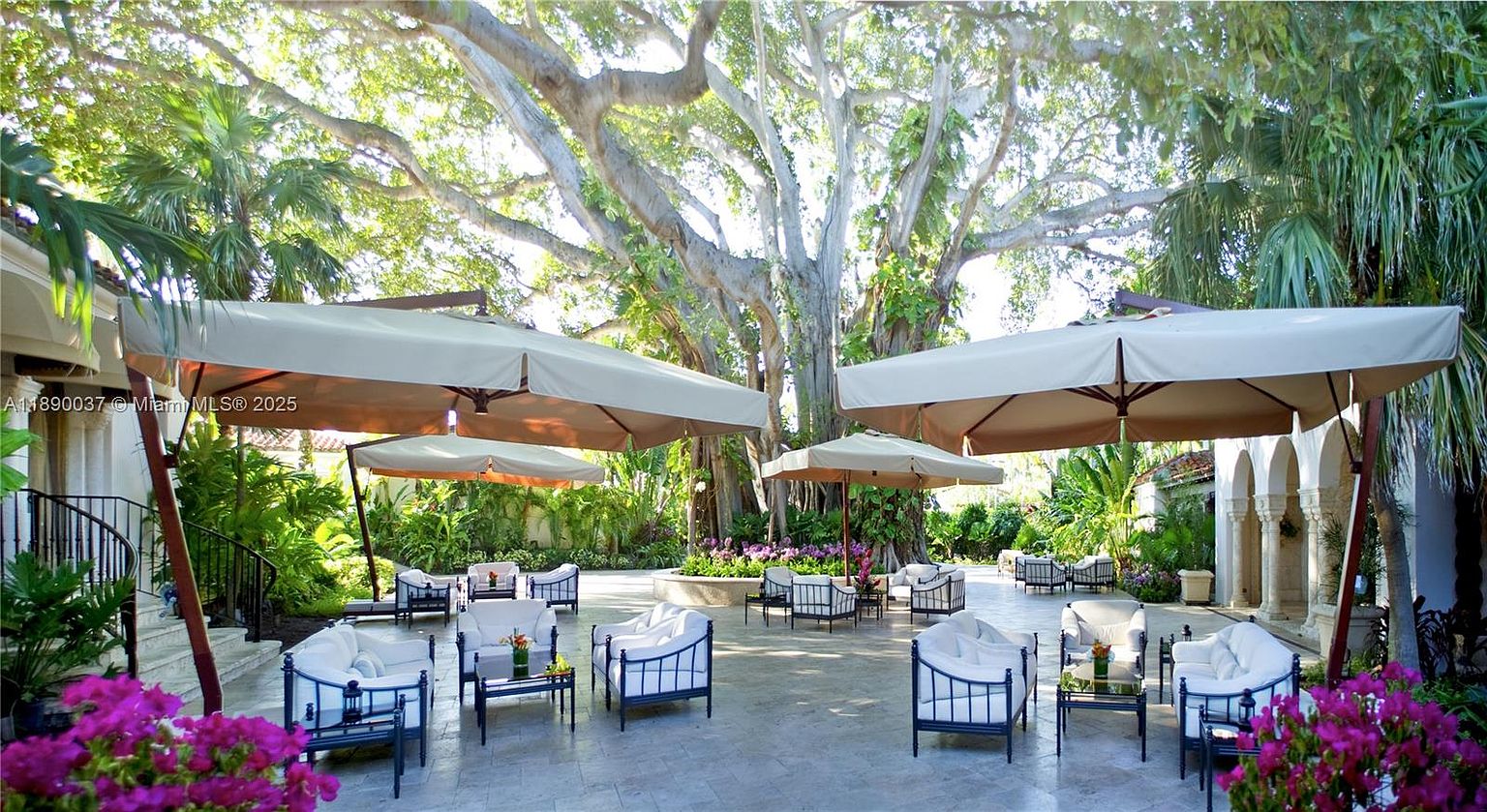This image showcases a luxurious outdoor patio area, furnished with elegant white cushioned seating and dark metal frames, arranged around small tables. Large, neutral-toned umbrellas provide shade, creating a comfortable and inviting space for relaxation or entertaining. The patio is surrounded by lush greenery and a mature tree, enhancing the serene and private atmosphere.
