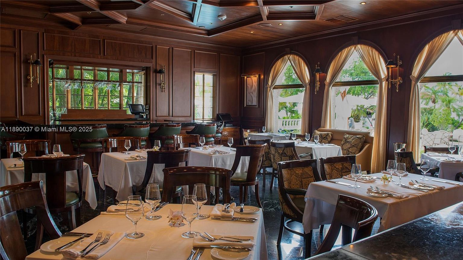 This is an interior shot of an elegant dining room, featuring dark wood paneling and arched windows draped with light curtains. Tables are set with white linens, silverware, and glassware, suggesting a fine dining experience. The room exudes a classic and sophisticated ambiance, perfect for a high-end restaurant or luxury home.