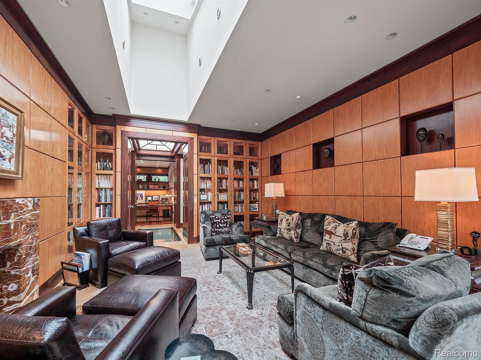 This is an interior shot of a luxurious living room featuring wood-paneled walls, built-in bookshelves, and comfortable seating arrangements. The room is well-lit with natural light from a skylight and features a mix of textures and patterns in the furniture and decor. The overall impression is one of sophistication and comfort, ideal for relaxation and entertaining.