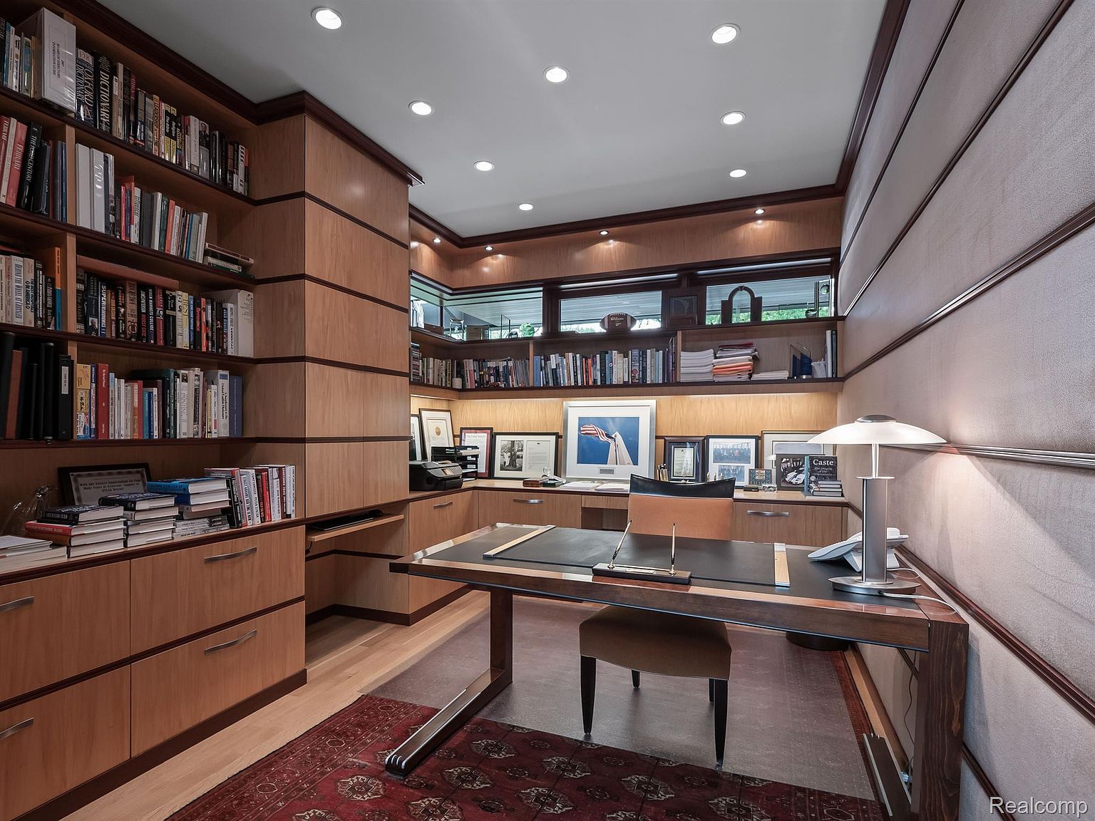 This is an interior shot of a well-appointed home office or study. The room features custom wood built-in shelving and cabinetry, a large desk with a leather top, and a comfortable chair. The lighting is provided by recessed ceiling lights and a desk lamp, creating a warm and inviting atmosphere.