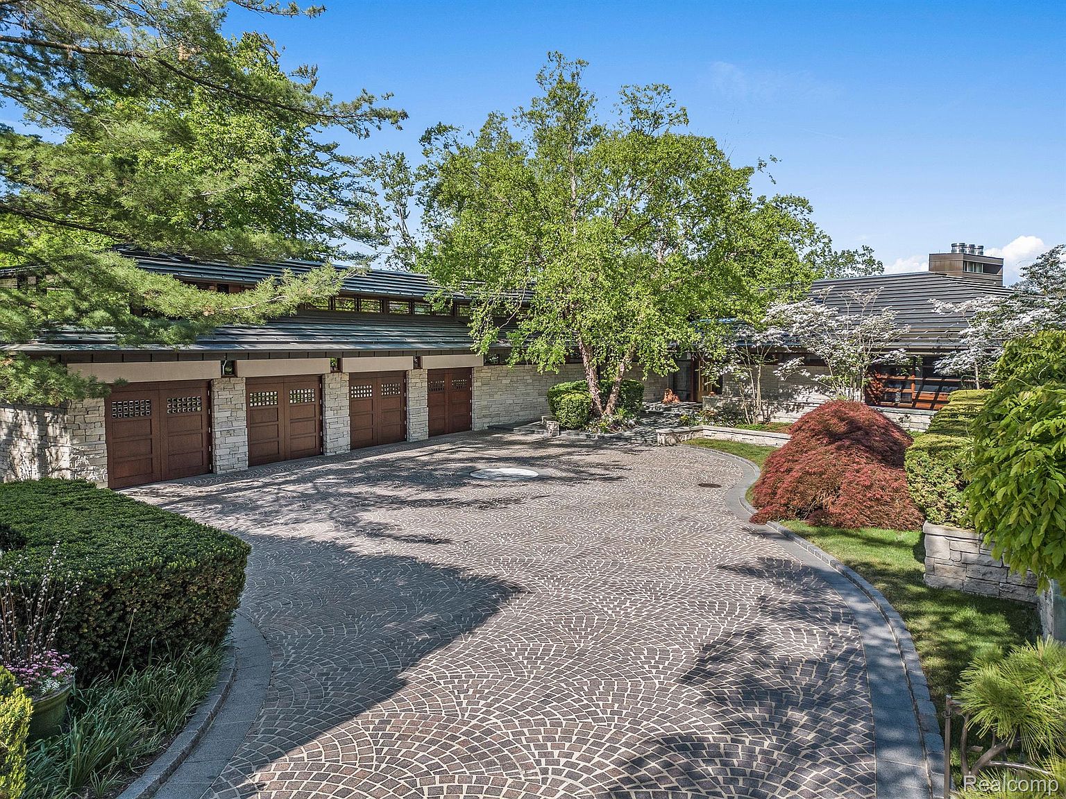 This image showcases the front exterior of a luxurious home with a unique architectural design. The property features a sprawling cobblestone driveway, a four-car garage with distinctive doors, and meticulously landscaped grounds with mature trees and manicured shrubbery. The overall impression is one of elegance and privacy.