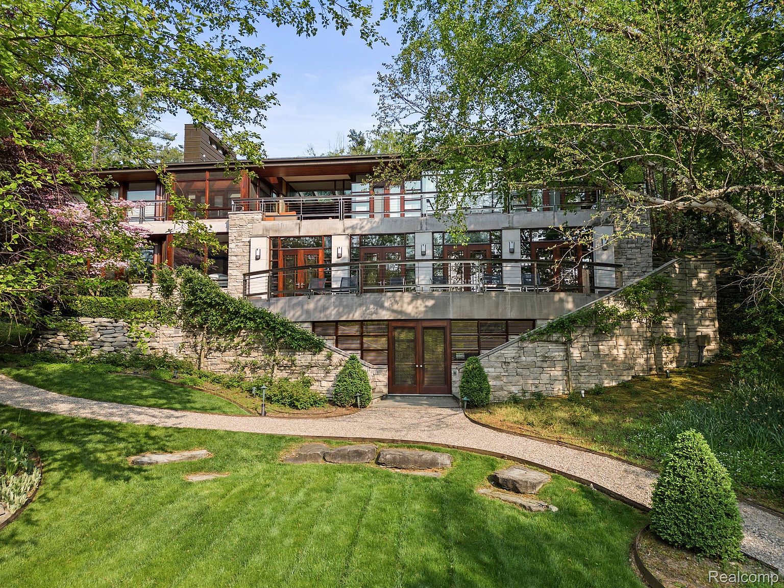 This is a front exterior view of a modern, multi-level home with a stone and concrete facade. The house features large windows and balconies, blending seamlessly with the surrounding natural landscape. A stone pathway leads to the entrance, surrounded by lush greenery and mature trees, creating a serene and private setting.