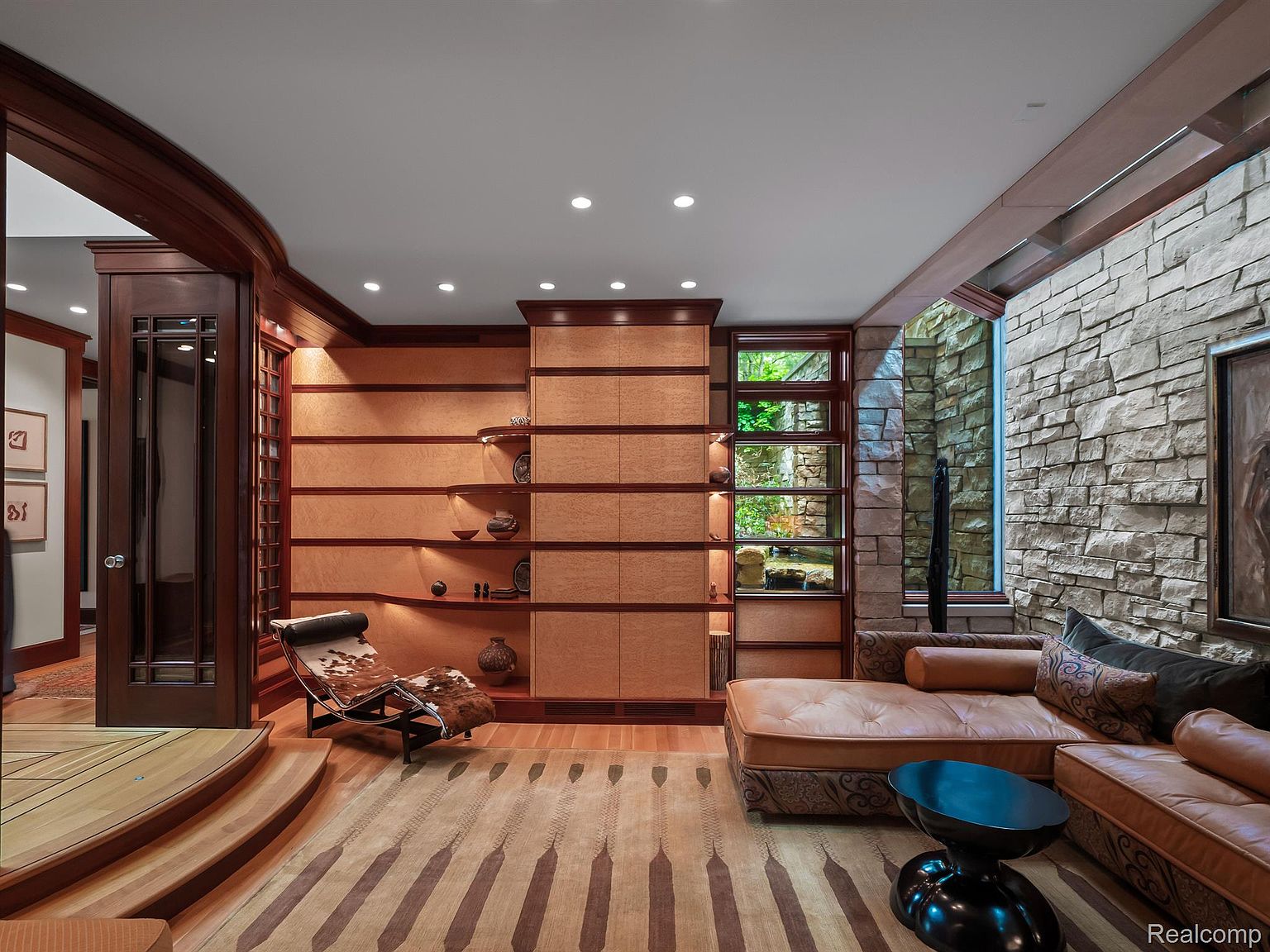 This interior shot showcases a luxurious living room with a unique architectural design. The room features a stone wall, built-in wooden shelving, and a comfortable leather sectional sofa. A distinctive rug and modern coffee table add to the room's sophisticated ambiance, creating an inviting and stylish space.