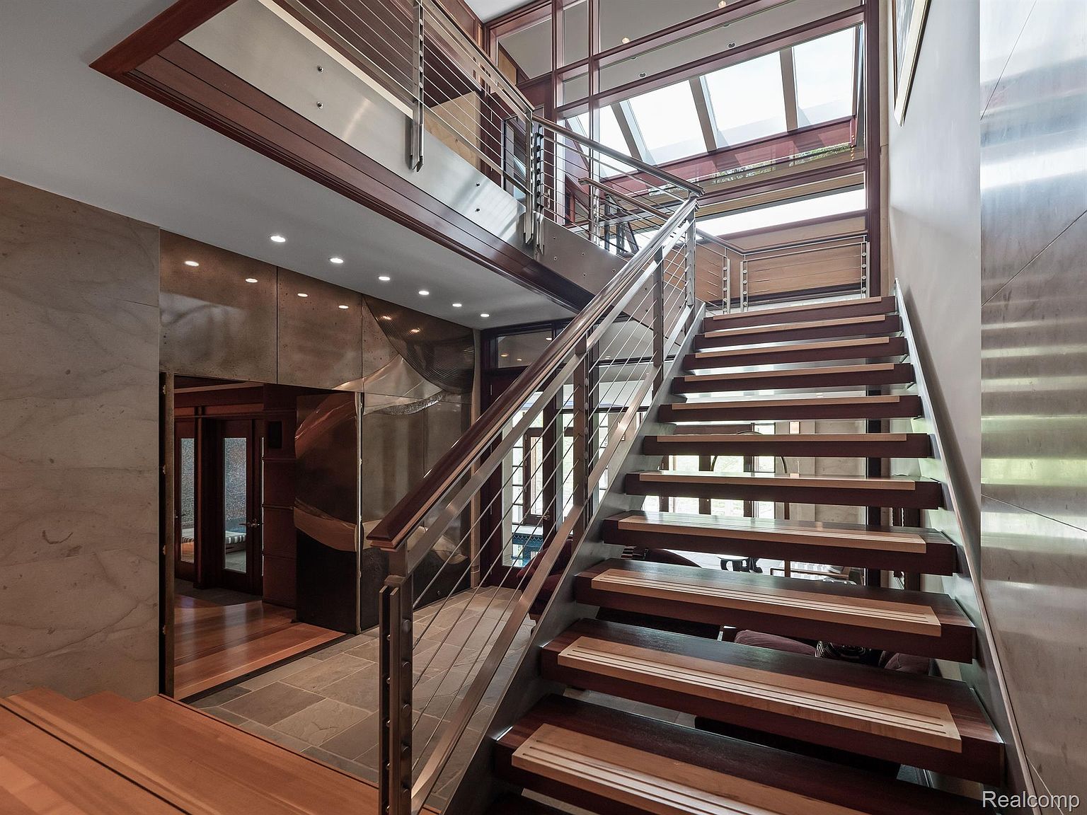 This interior shot showcases a modern hallway and staircase. The staircase features wooden steps with metal railings, leading to an upper level with a glass-enclosed area. The hallway is adorned with unique wall finishes and recessed lighting, creating a sophisticated and well-lit transition space.