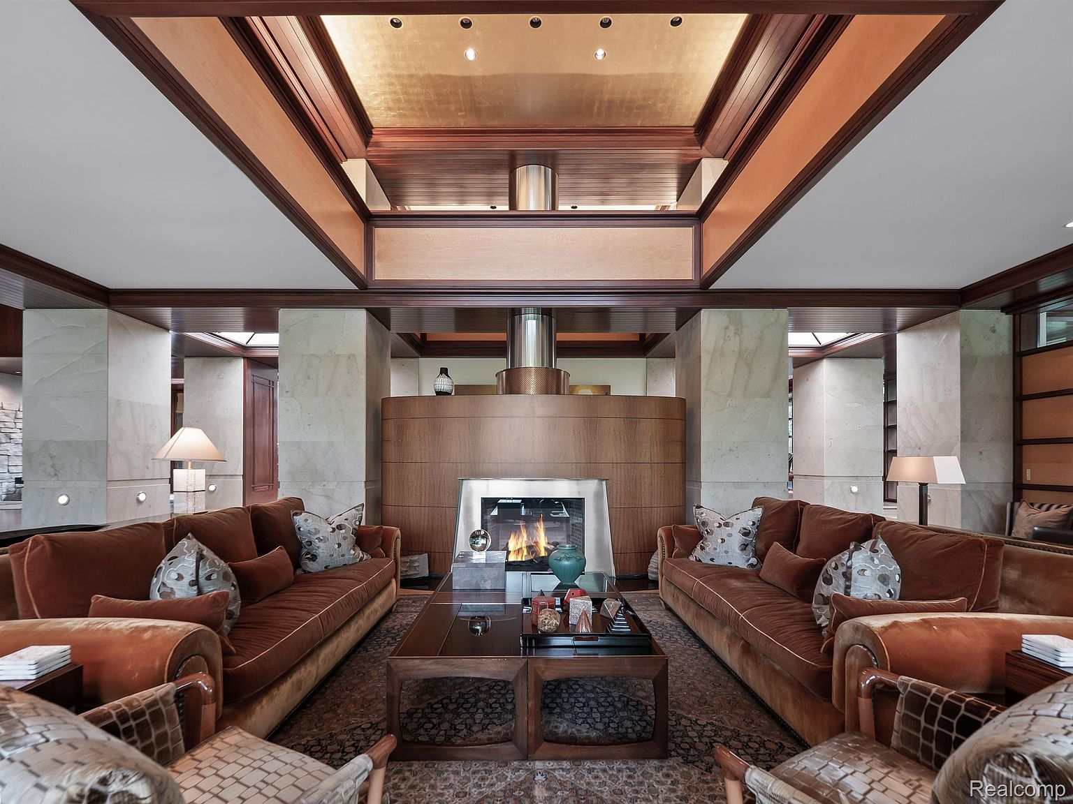 This is an interior shot of a luxurious living room featuring two large, brown velvet sofas facing each other, flanking a dark wood coffee table. The room is centered around a modern fireplace with a stainless steel chimney, and the ceiling is a complex design of wood and recessed lighting. The overall impression is one of warmth, sophistication, and high-end design.