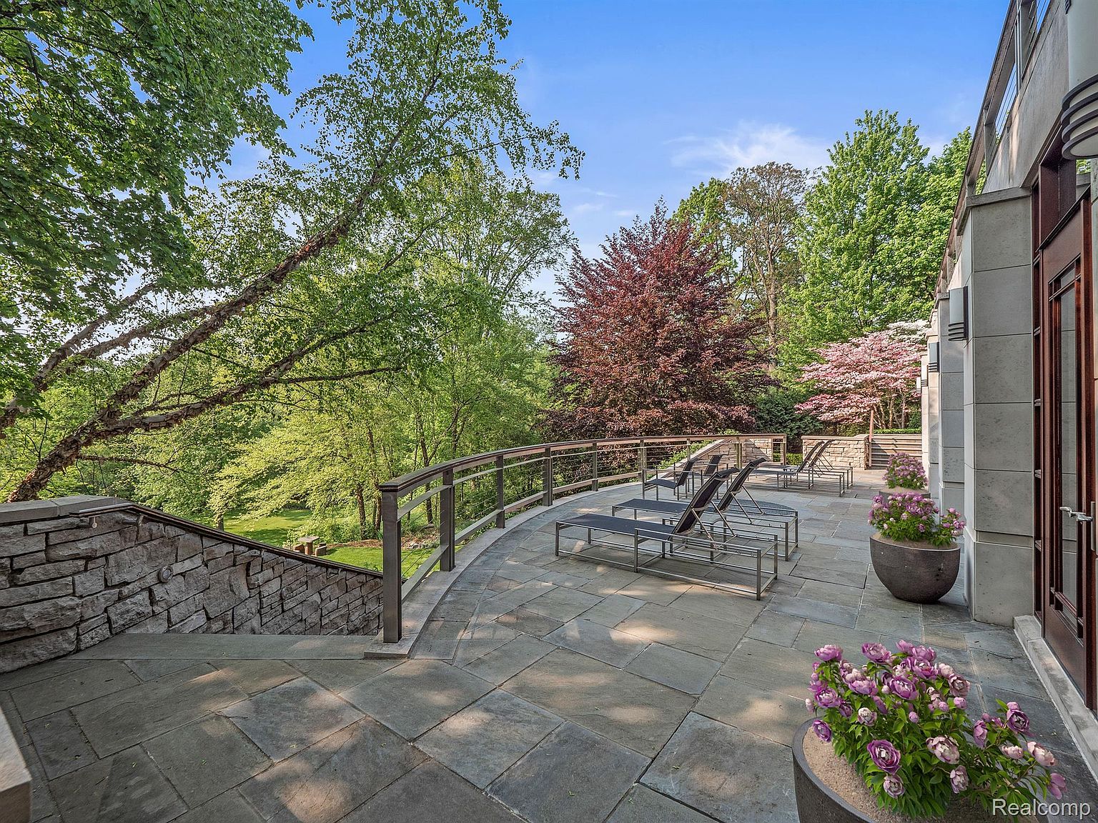 This image showcases a luxurious patio or deck area, featuring stone flooring and a metal railing that overlooks a lush, green landscape. Several lounge chairs are neatly arranged, suggesting a relaxing outdoor space. Potted flowers add a touch of color and elegance, enhancing the overall appeal of this outdoor living area.