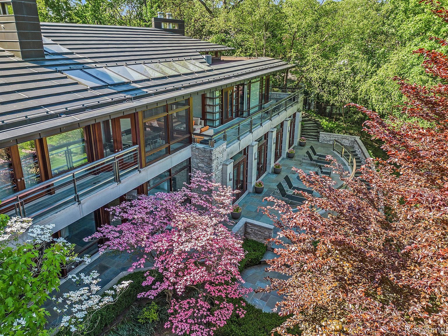 This aerial view showcases a modern home with a striking roofline, expansive windows, and multiple balconies. The property is surrounded by lush landscaping, including flowering trees and manicured gardens, creating a serene and private setting. The architectural design emphasizes a seamless integration of indoor and outdoor living spaces.