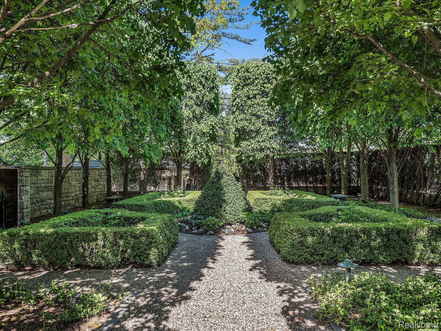This image showcases a meticulously maintained garden with symmetrical hedges, gravel pathways, and mature trees providing shade. The garden's design creates a sense of tranquility and order, enhancing the property's curb appeal. The perspective is from the entrance of the garden, drawing the viewer into the space.
