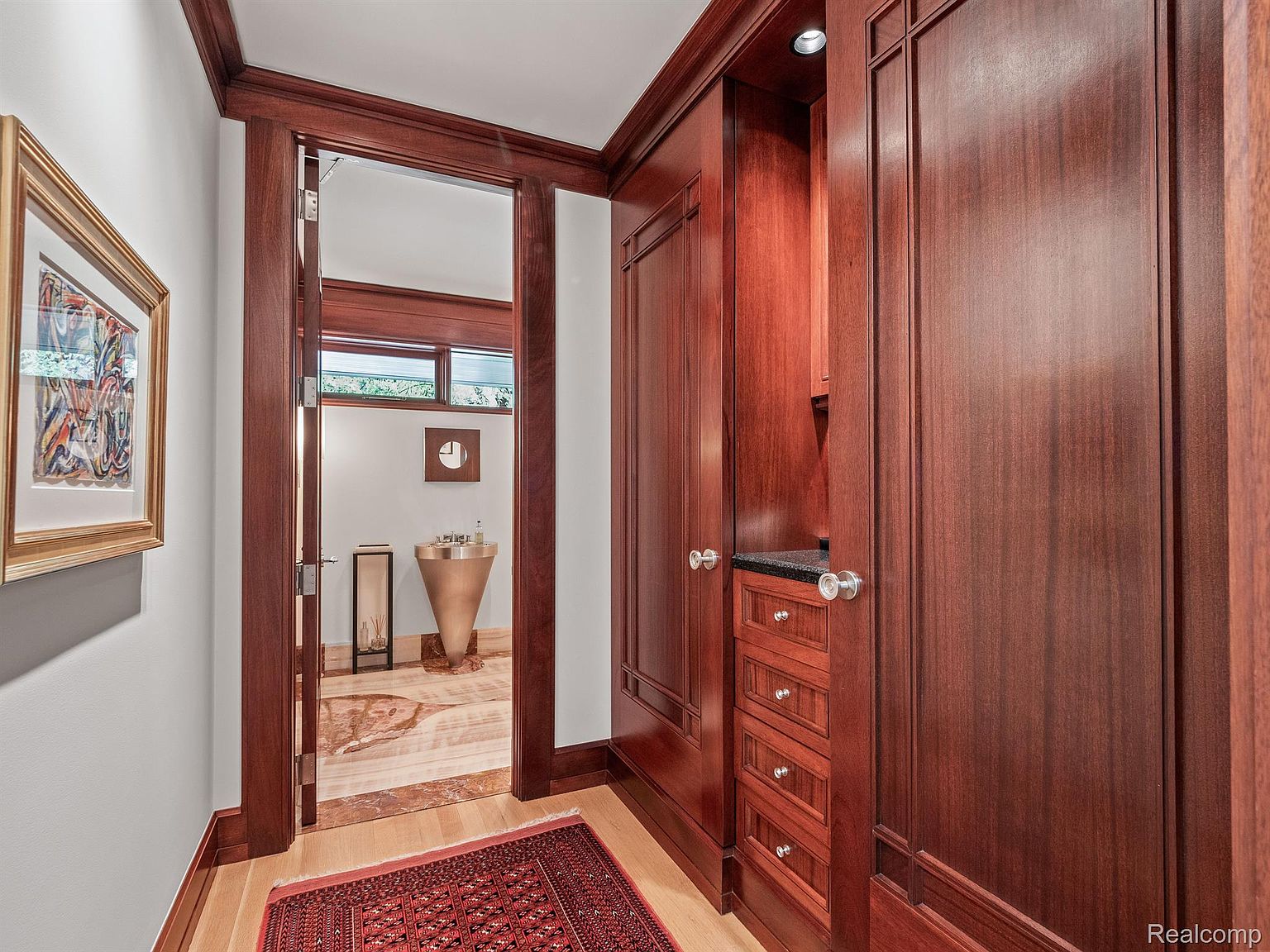 This interior shot showcases a hallway with rich, dark wood paneling and trim, creating a luxurious and sophisticated atmosphere. A doorway leads to a bathroom with a unique, cone-shaped sink. A red oriental rug adds a pop of color and warmth to the hardwood flooring, enhancing the overall elegance of the space.