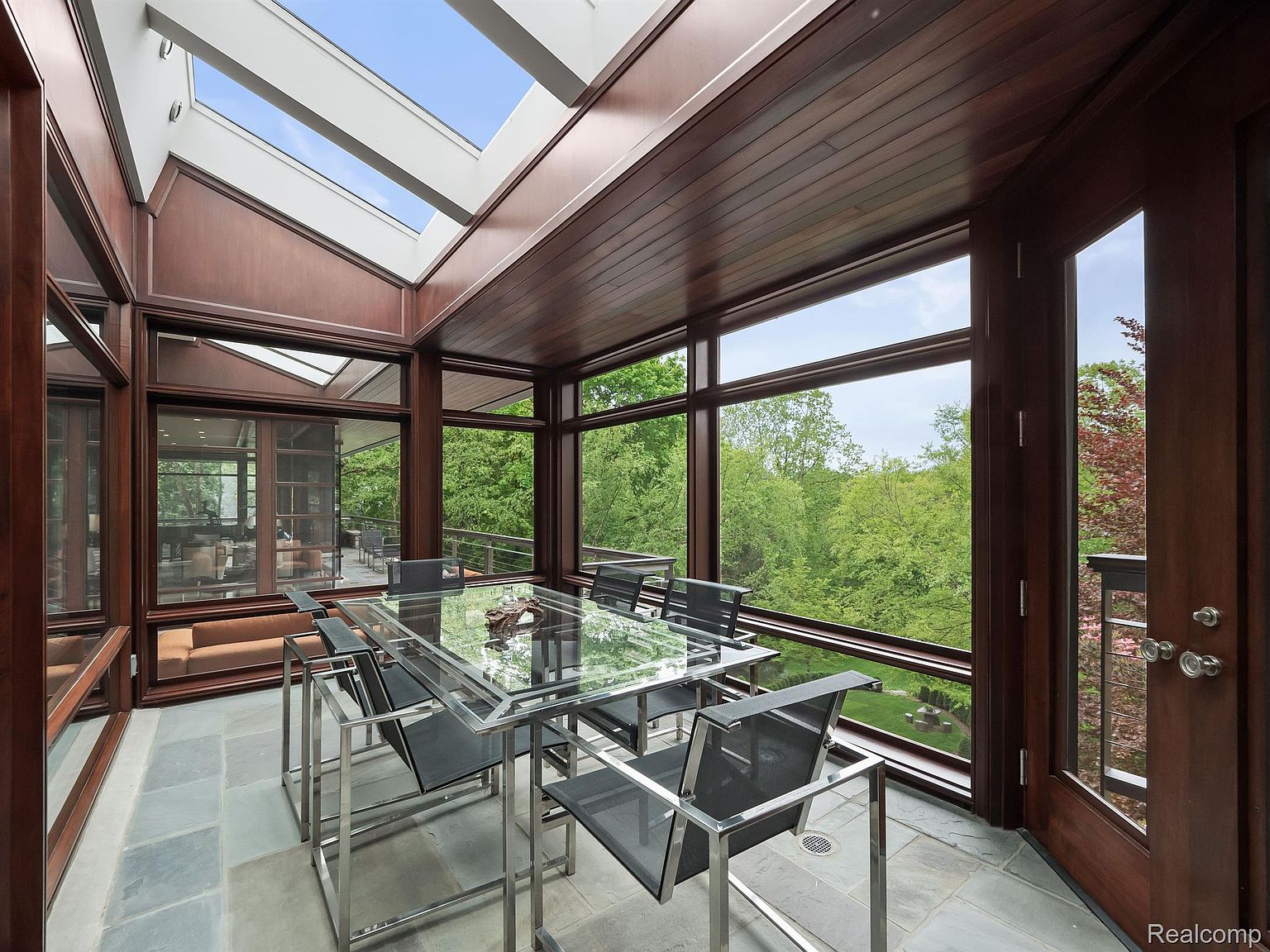 This sunroom features a glass-topped dining table with modern chairs, surrounded by large windows offering views of lush greenery. The room has a wood-paneled ceiling with skylights, and the floor is tiled with large stone tiles. The space is bright and airy, creating a serene dining environment.