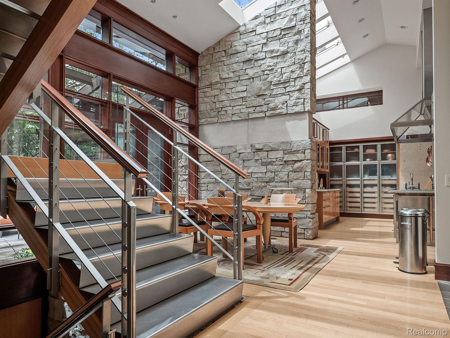 This interior shot showcases a modern home's hallway and staircase, featuring stainless steel railings and light wood steps. A stone accent wall adds texture and visual interest, complemented by natural light streaming through skylights. The open layout connects the hallway to a dining area and kitchen, creating a spacious and inviting atmosphere.