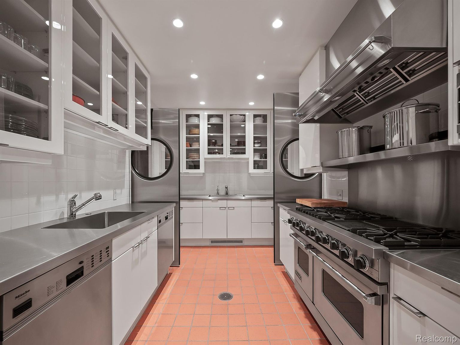 This is a well-lit, modern kitchen featuring stainless steel countertops and appliances, including a Miele dishwasher and a professional-grade gas range with a large hood. White cabinetry with glass fronts provides ample storage, and the terracotta tile flooring adds a touch of warmth to the space. The perspective is a straight-on shot, emphasizing the depth and symmetry of the kitchen layout.