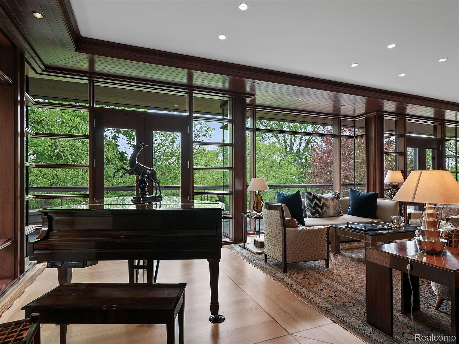 This is an elegant living room featuring floor-to-ceiling windows that offer a view of lush greenery. A grand piano sits prominently in the room, complemented by sophisticated furniture and a patterned area rug. The dark wood trim and recessed lighting add to the room's luxurious ambiance.